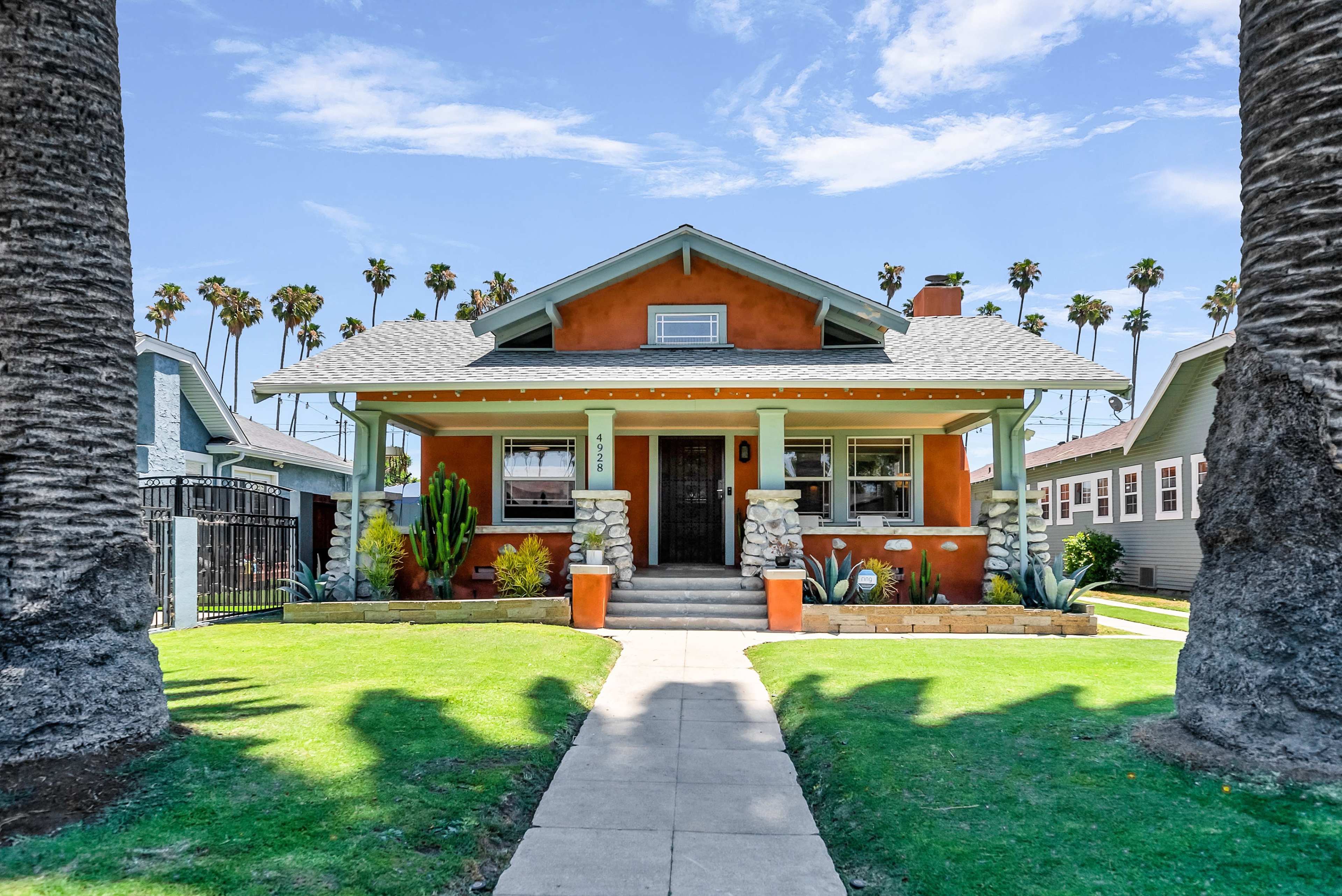 A vibrant orange bungalow with stone accents sits under tall palm trees, featuring a front path lined with greenery.
