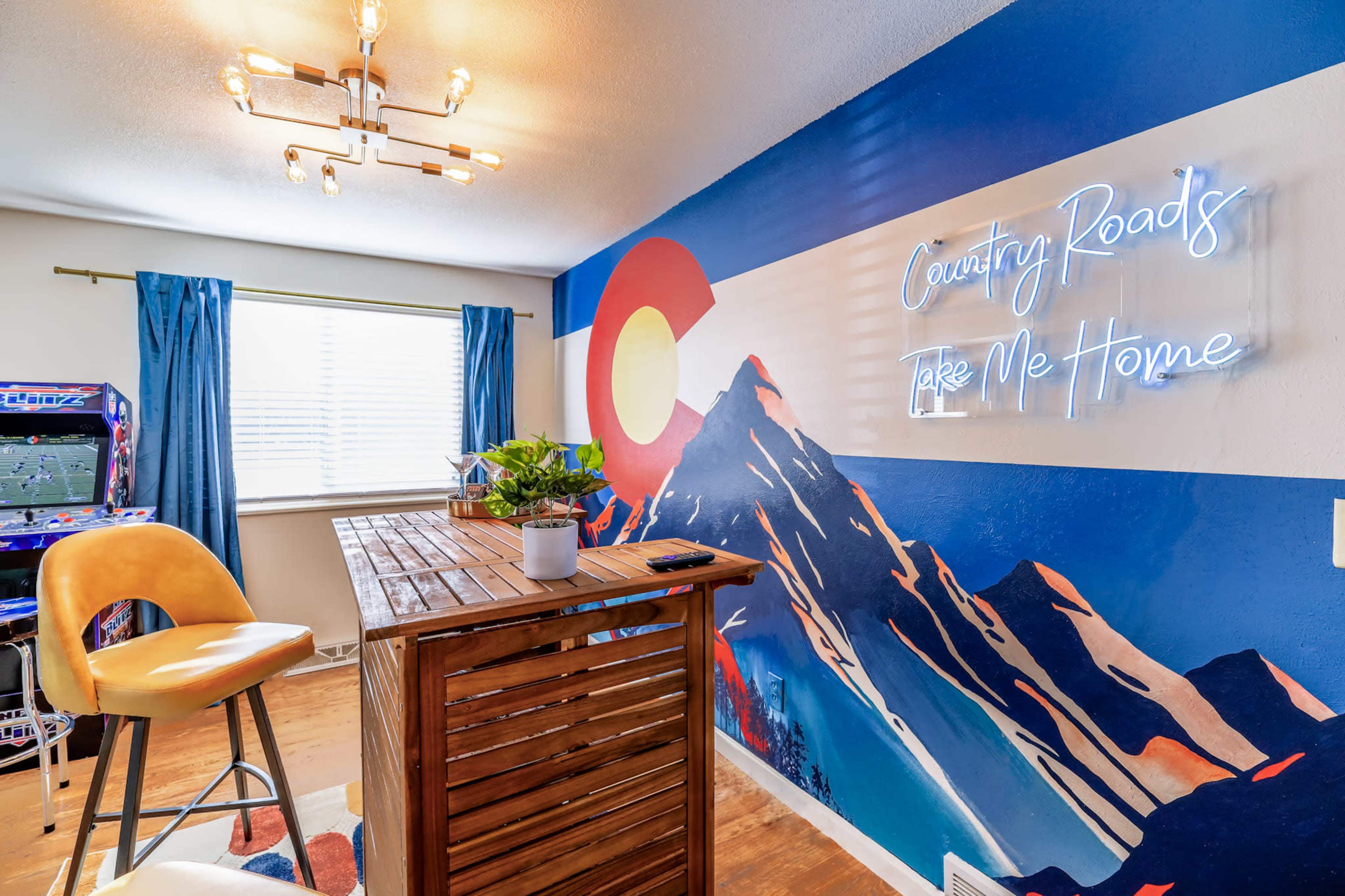 The room features a wooden bar table against a mural of mountains and a Colorado-themed wall, illuminated by a neon sign.