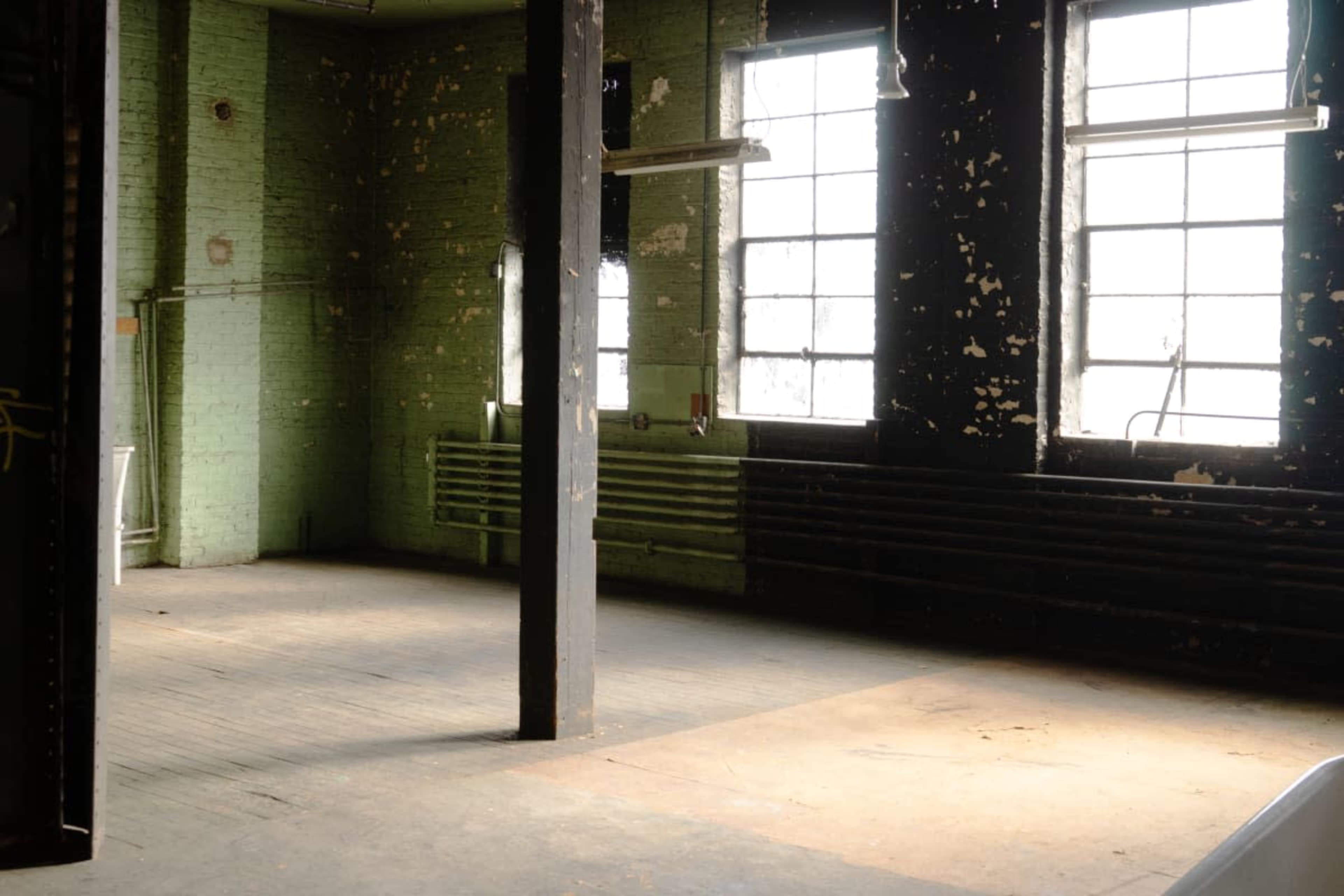 The image shows an empty, worn interior space with peeling paint on the walls, exposed pipes, and large windows allowing natural light to enter.