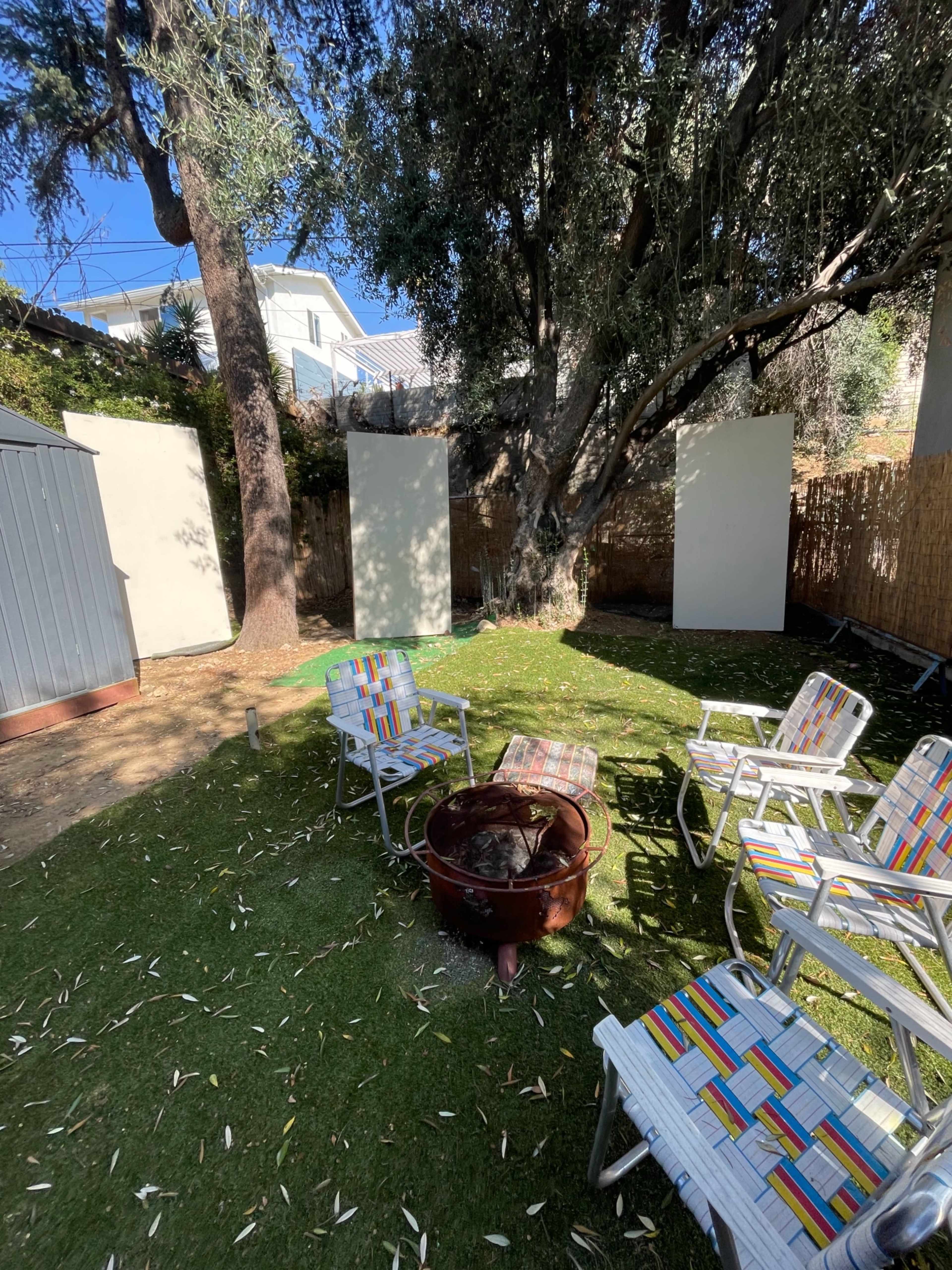 A grassy backyard features several striped chairs arranged around a fire pit, with white walls standing in the background.
