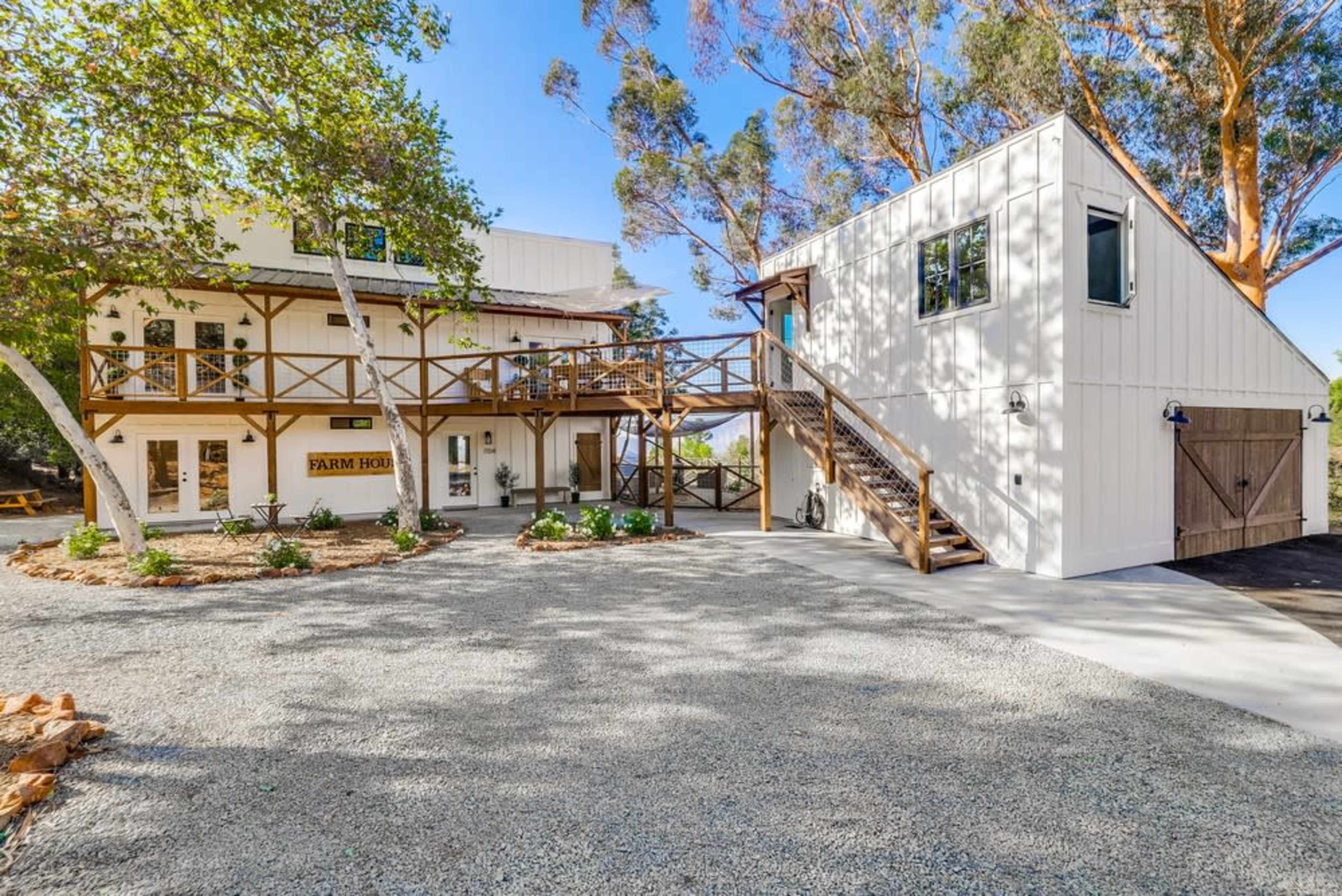 A two-story farmhouse with a wooden deck and stairs, surrounded by gravel and greenery.