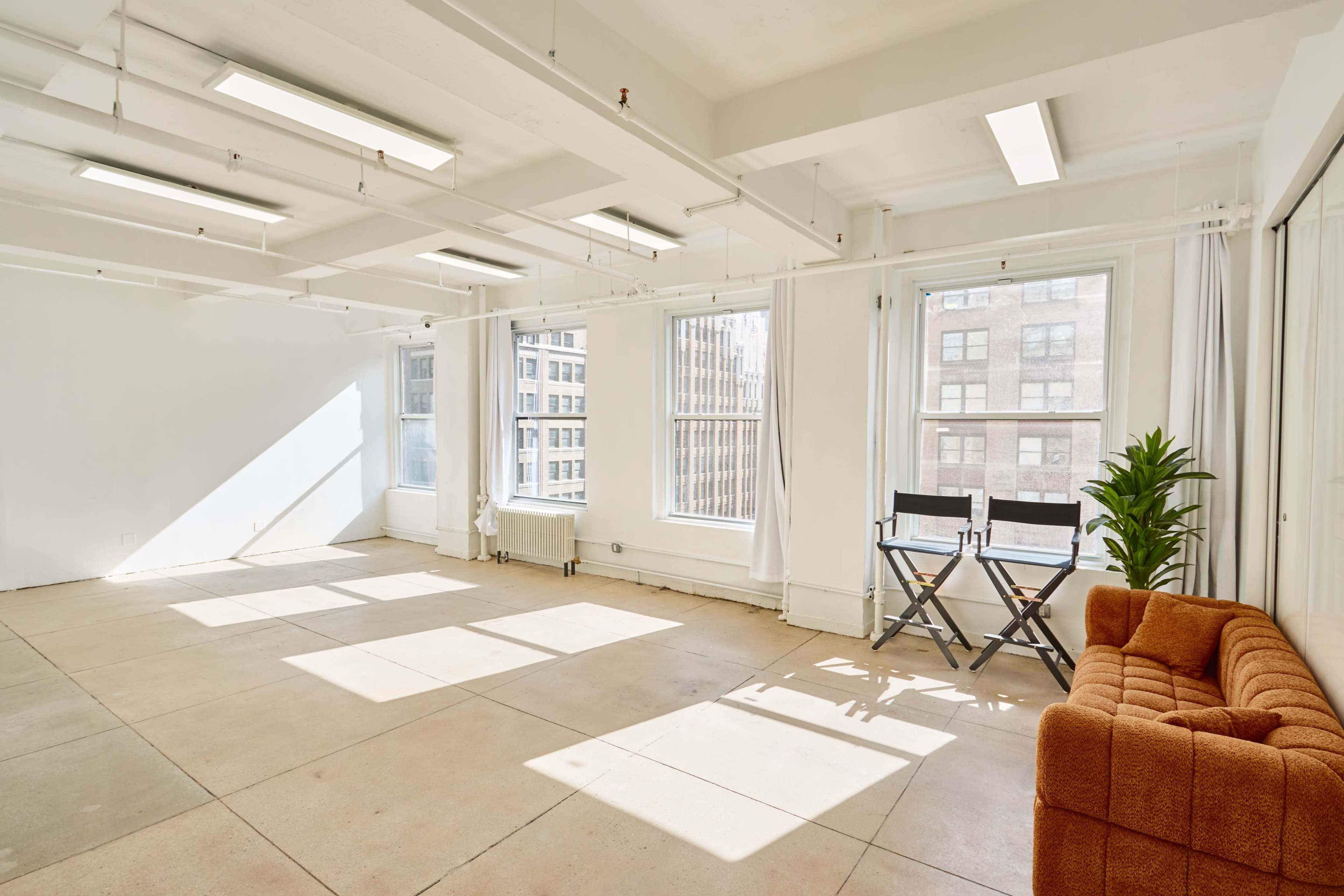 A brightly lit room with large windows features a couch, folding chairs, and light-colored walls.
