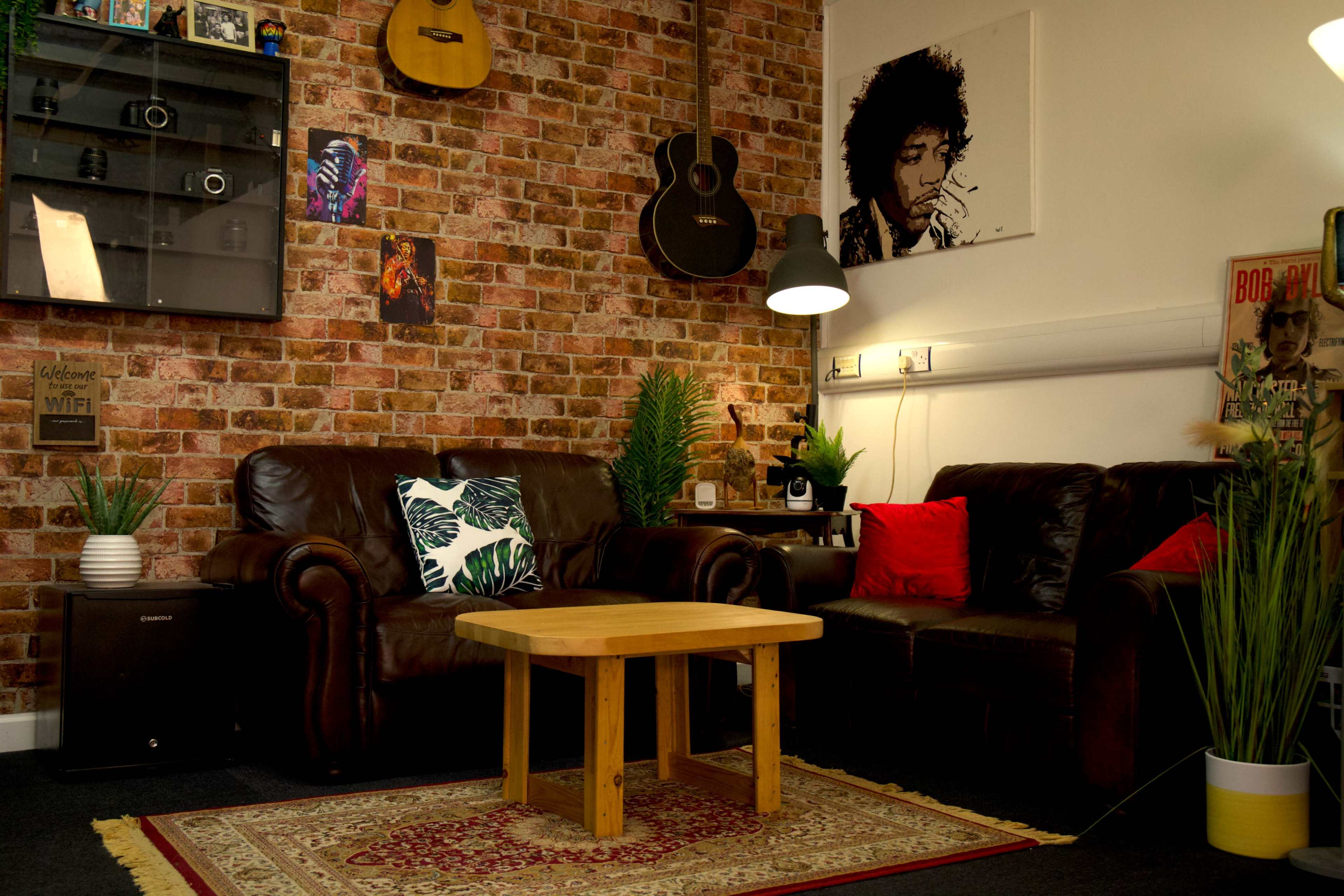 The image shows a cozy living room with brown leather sofas, a wooden coffee table, decorative plants, and a brick-patterned wall featuring various artworks and musical instruments.