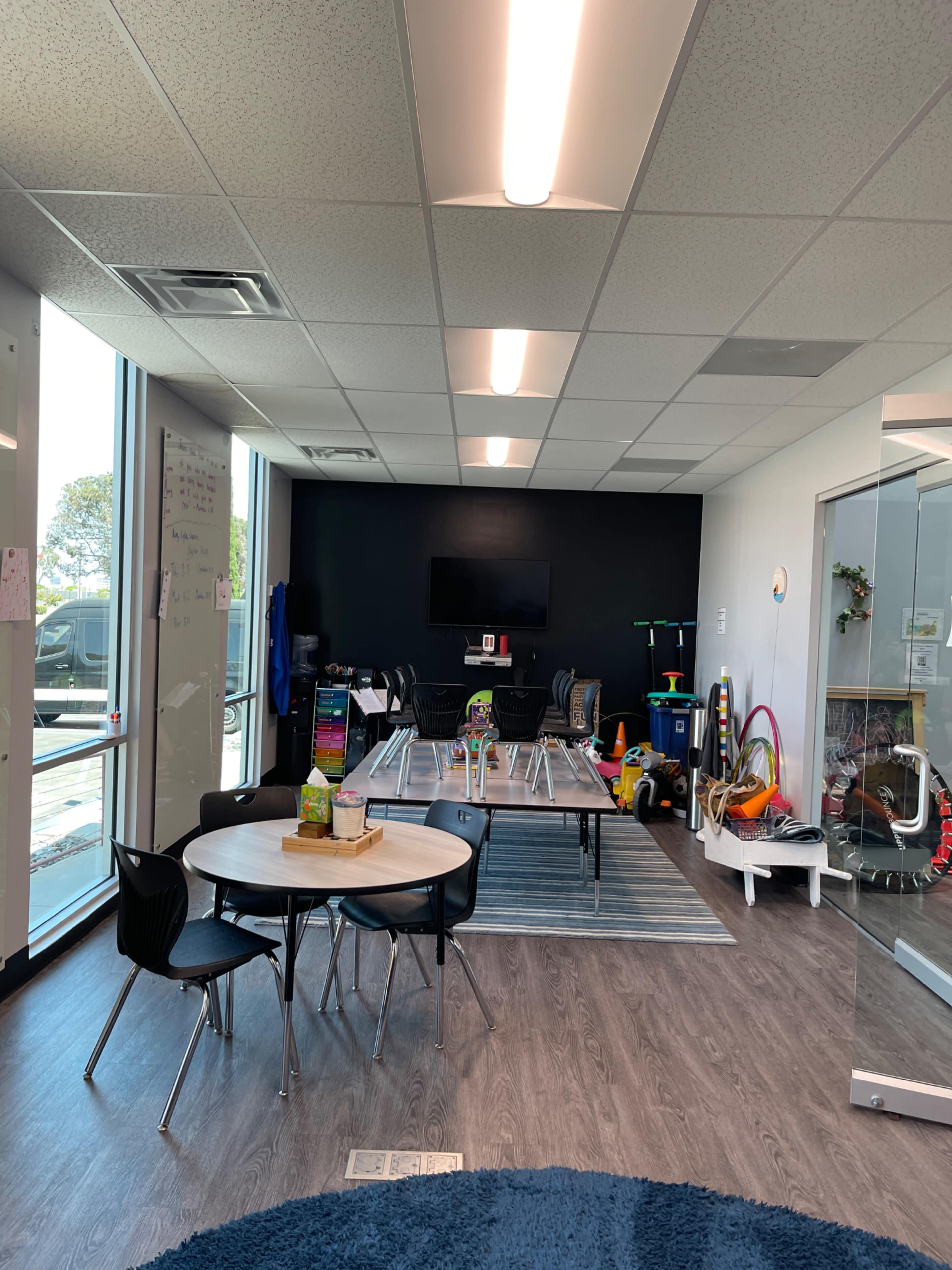 The image shows a classroom or activity space with tables and chairs arranged near a wall-mounted television, surrounded by various educational materials and toys.