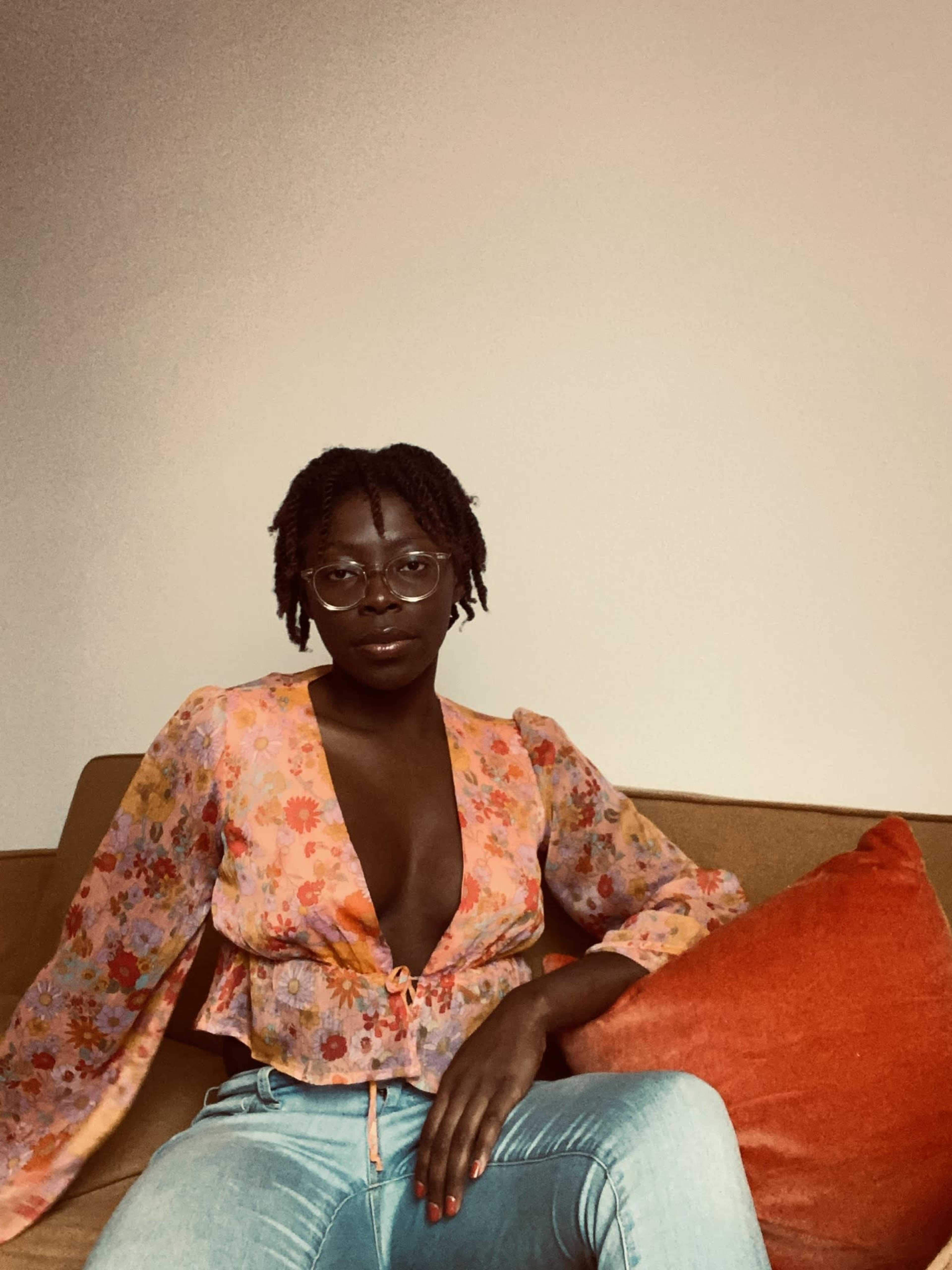 A person wearing a floral blouse and glasses sits on a couch next to an orange pillow.