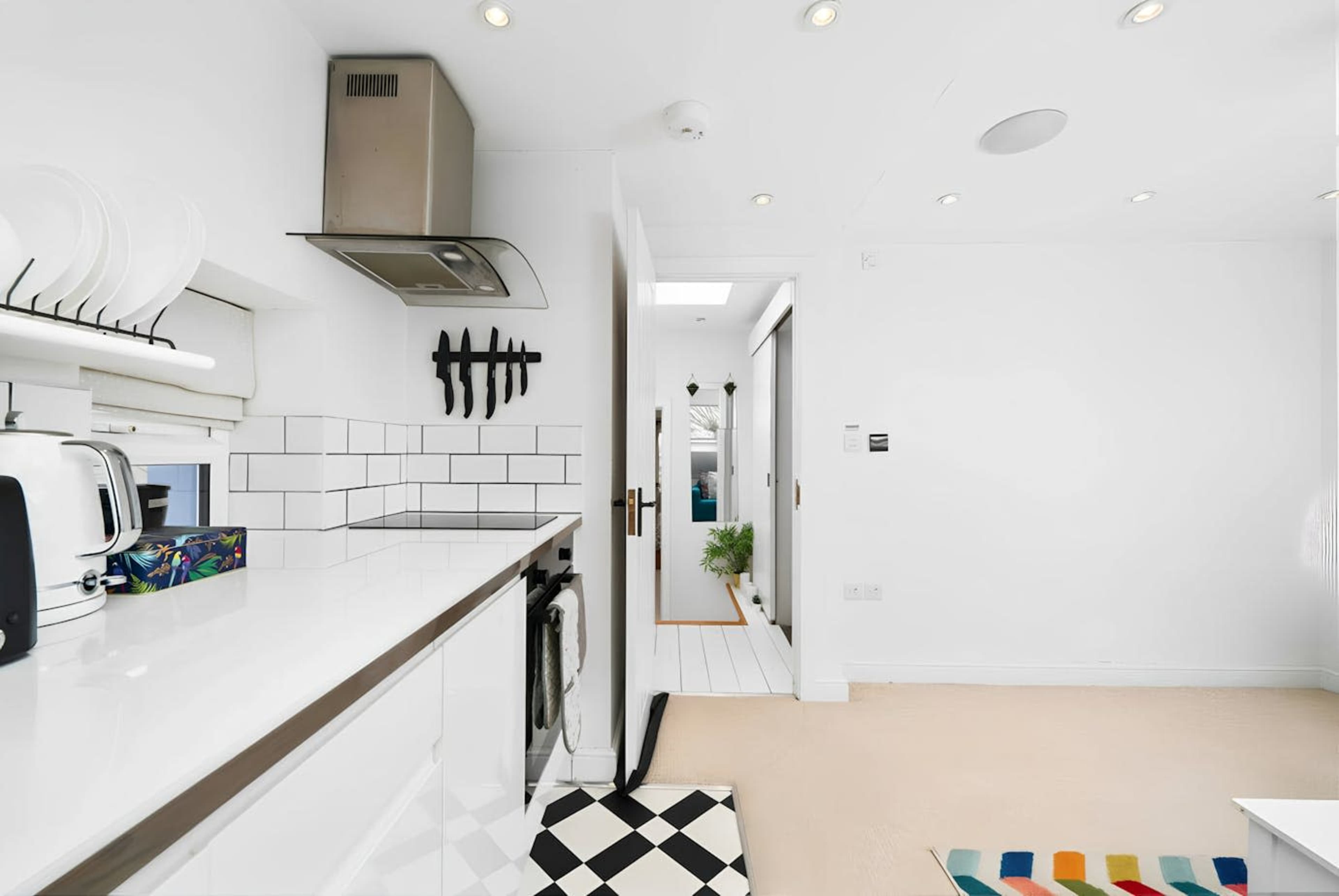 The image shows a bright, modern kitchen with white cabinetry, a black-and-white checkered floor, and a view of a hallway leading to another room.