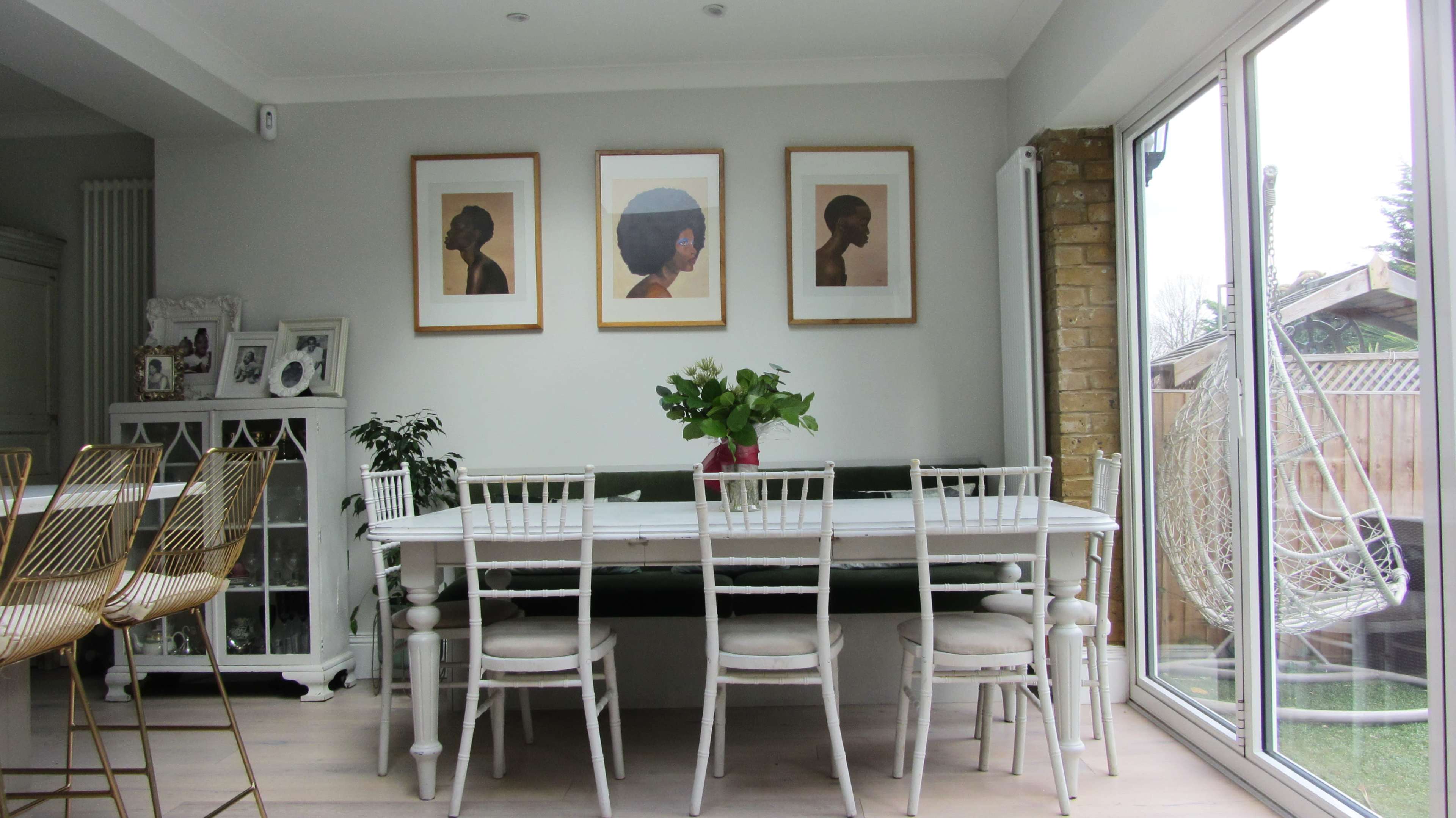 A bright dining area features a long white table surrounded by chairs, with framed portraits on the wall and a window that opens to a garden view.