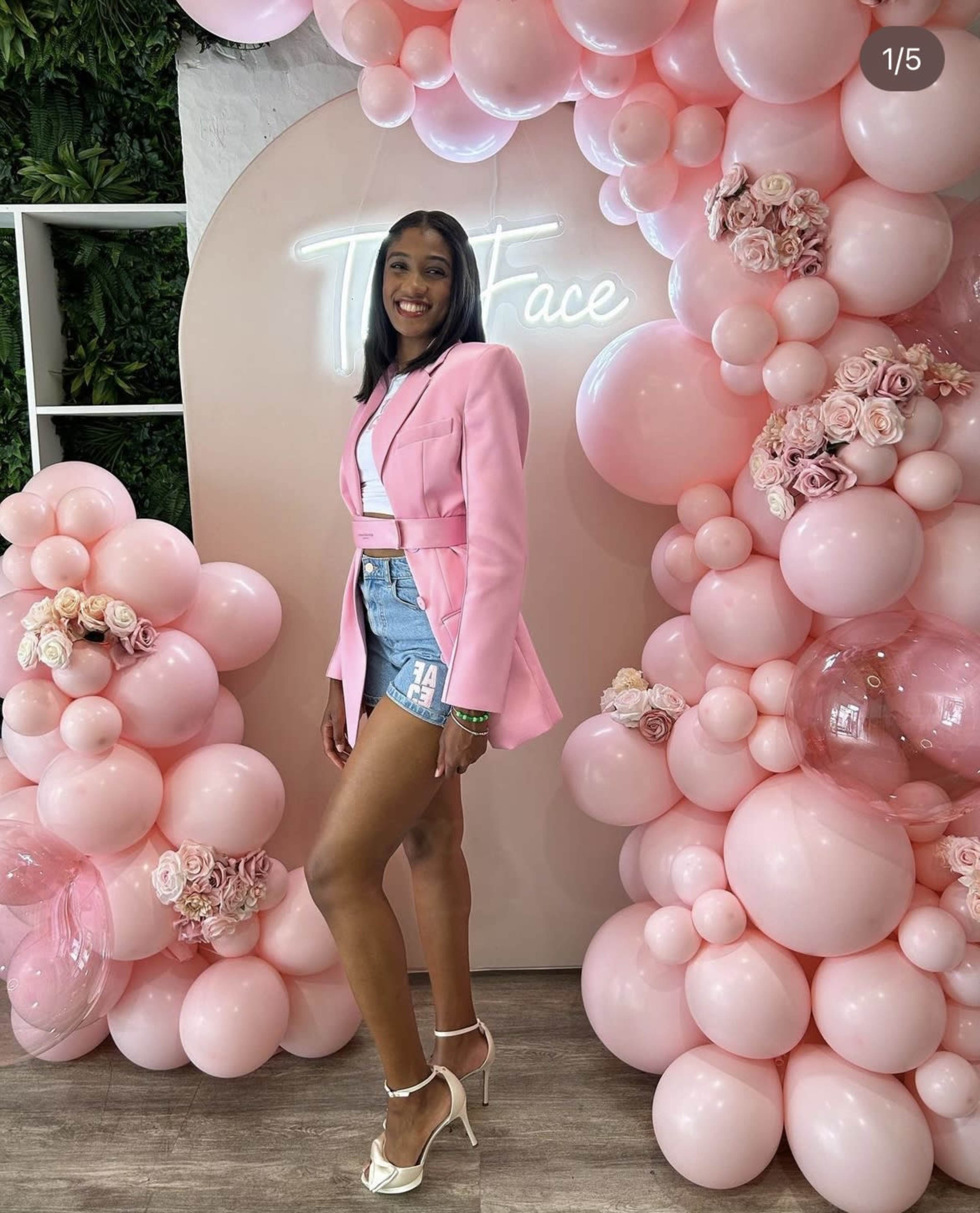 A woman stands in front of a backdrop adorned with pink balloons and flowers, wearing a pink blazer and denim shorts.
