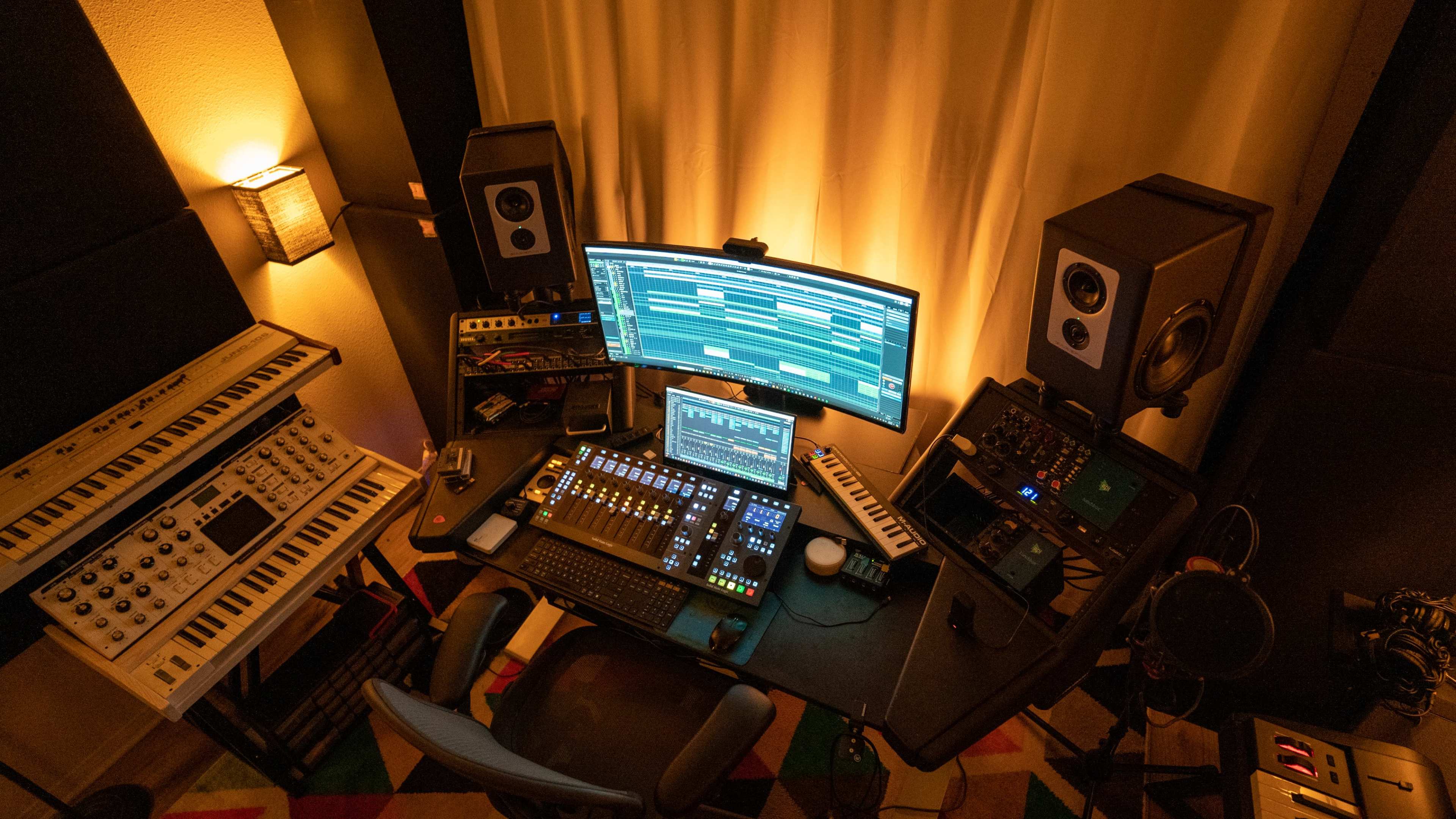 A music production studio featuring a large monitor, audio speakers, a mixing console, and various electronic instruments arranged around a desk.