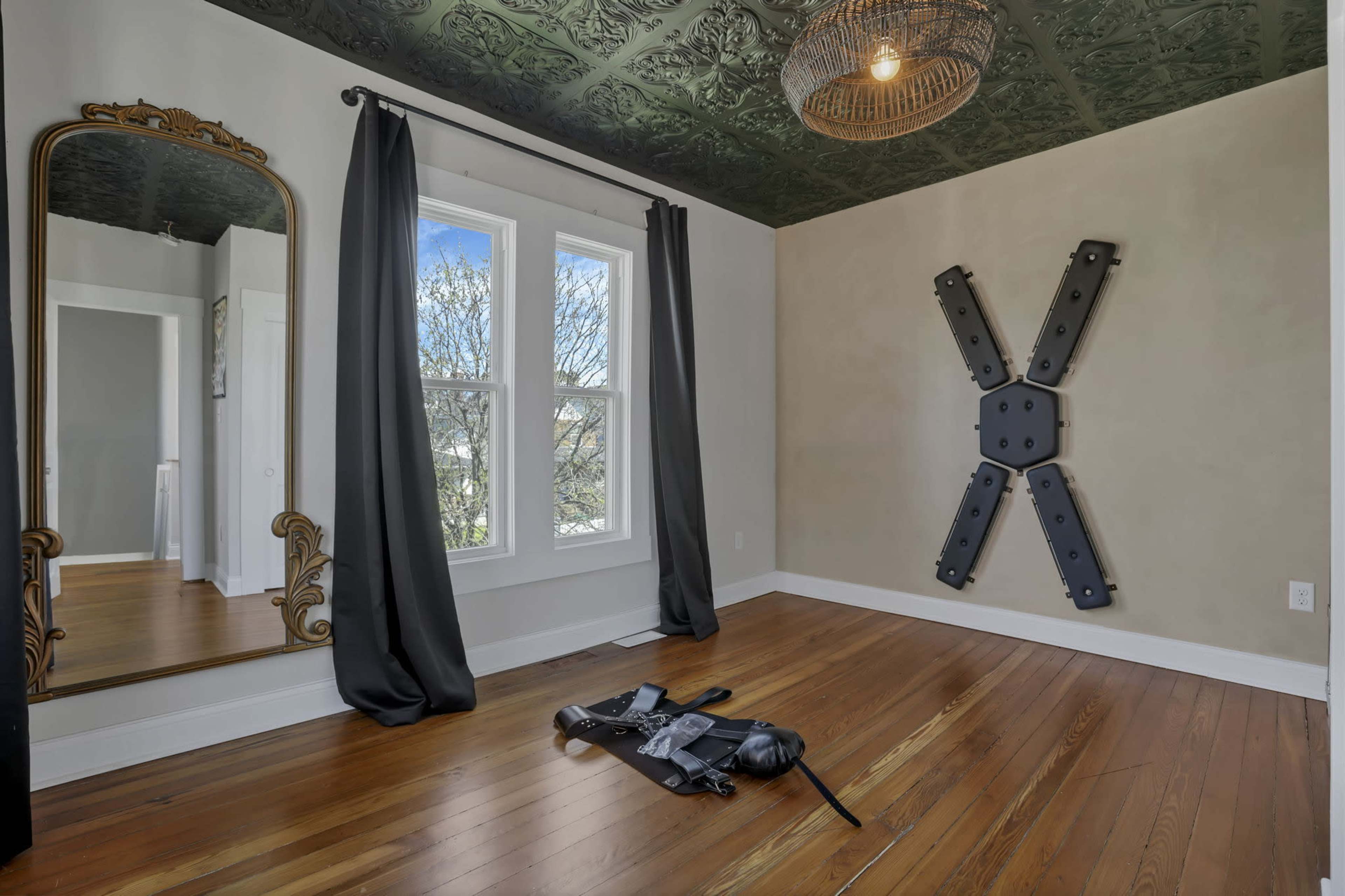 A room features a mirror with an ornate frame, large windows with dark curtains, a metal wall decoration, and wooden flooring.
