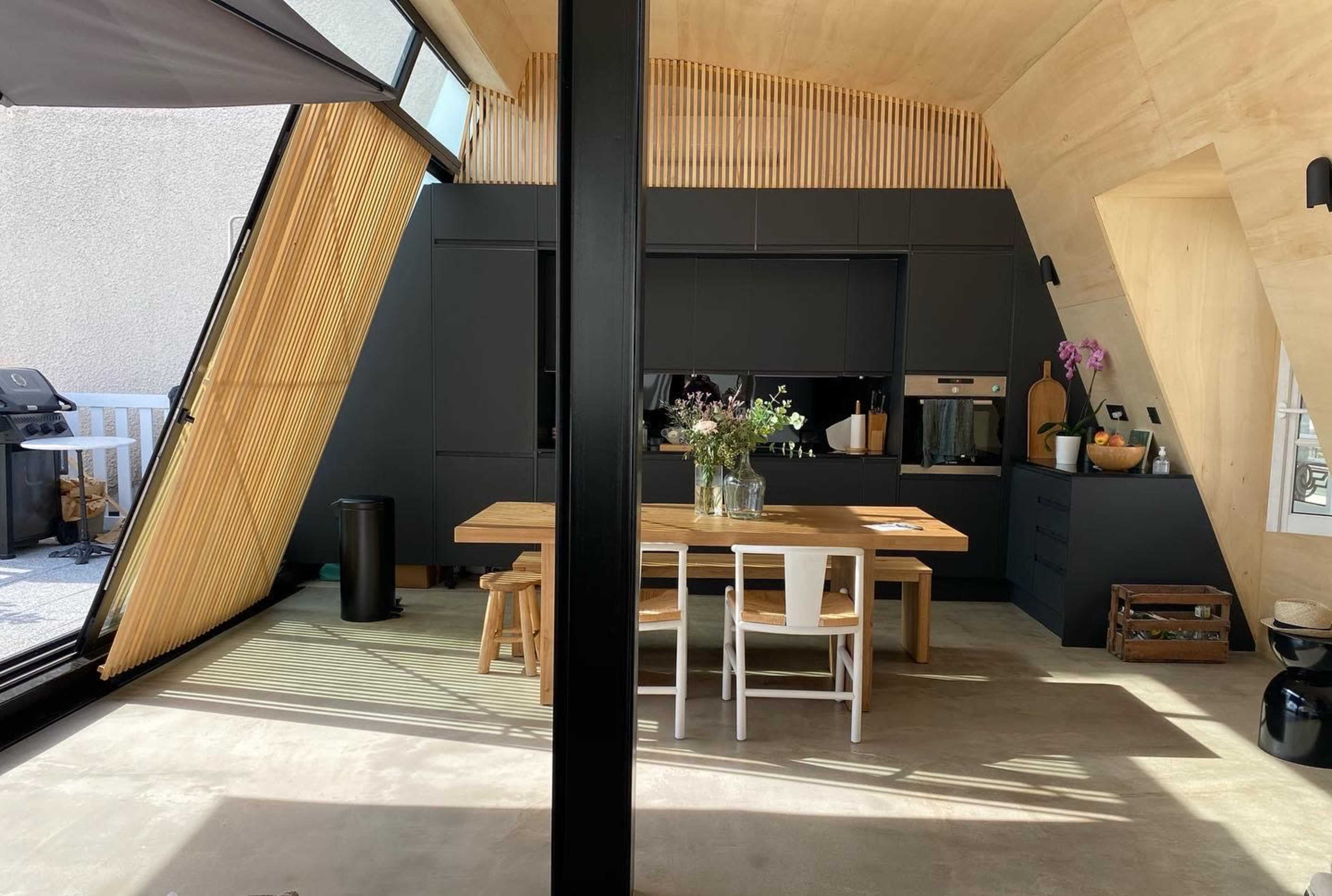 A modern kitchen with an angled wooden ceiling, a large dining table, and sleek black cabinets, illuminated by natural light from large windows.
