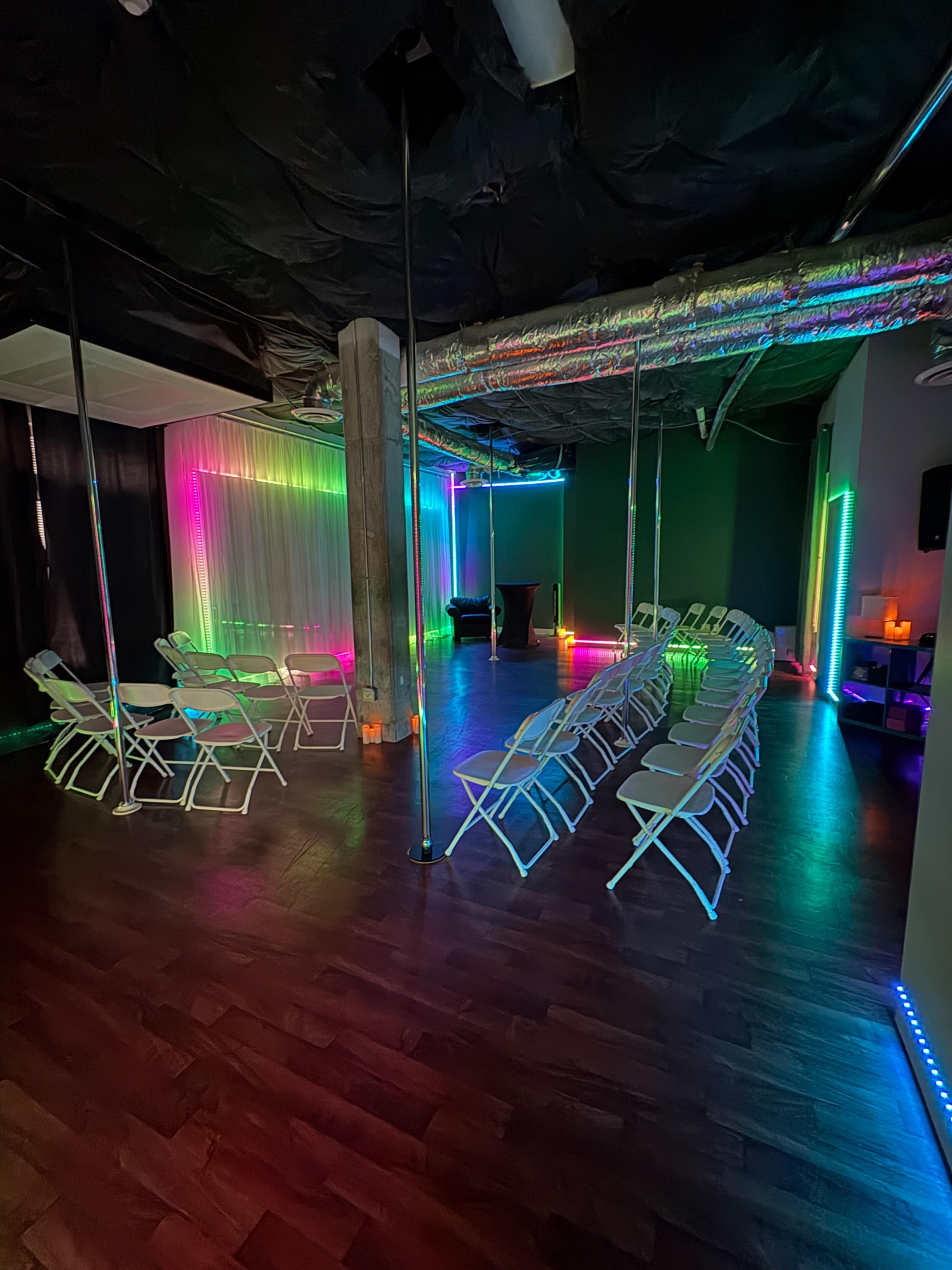 The image shows a room set up for an event, featuring rows of folding chairs, colored lights along the walls, and poles in the center.