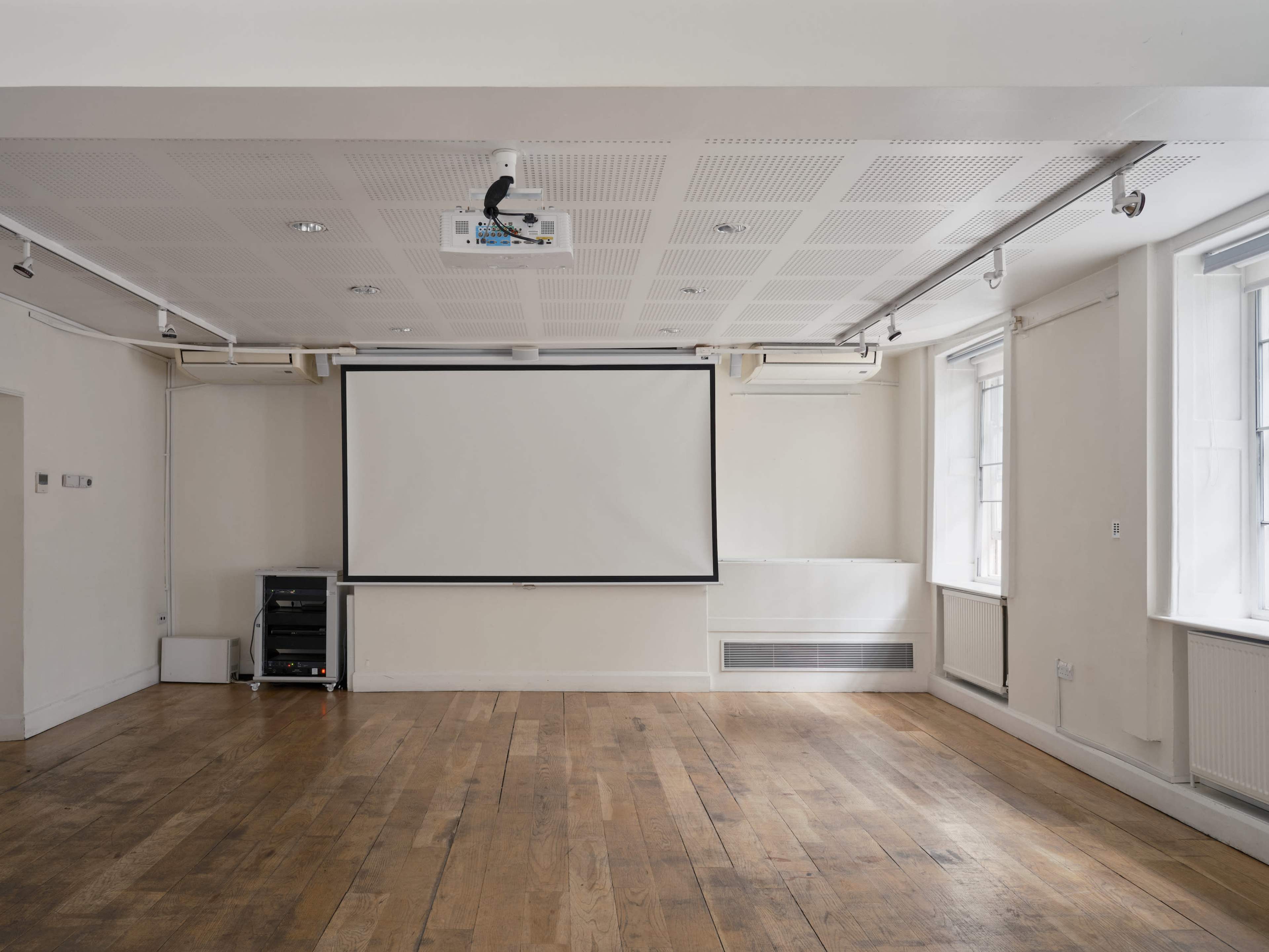 The image shows a spacious, empty room with wooden floors and a projector screen mounted on the far wall.