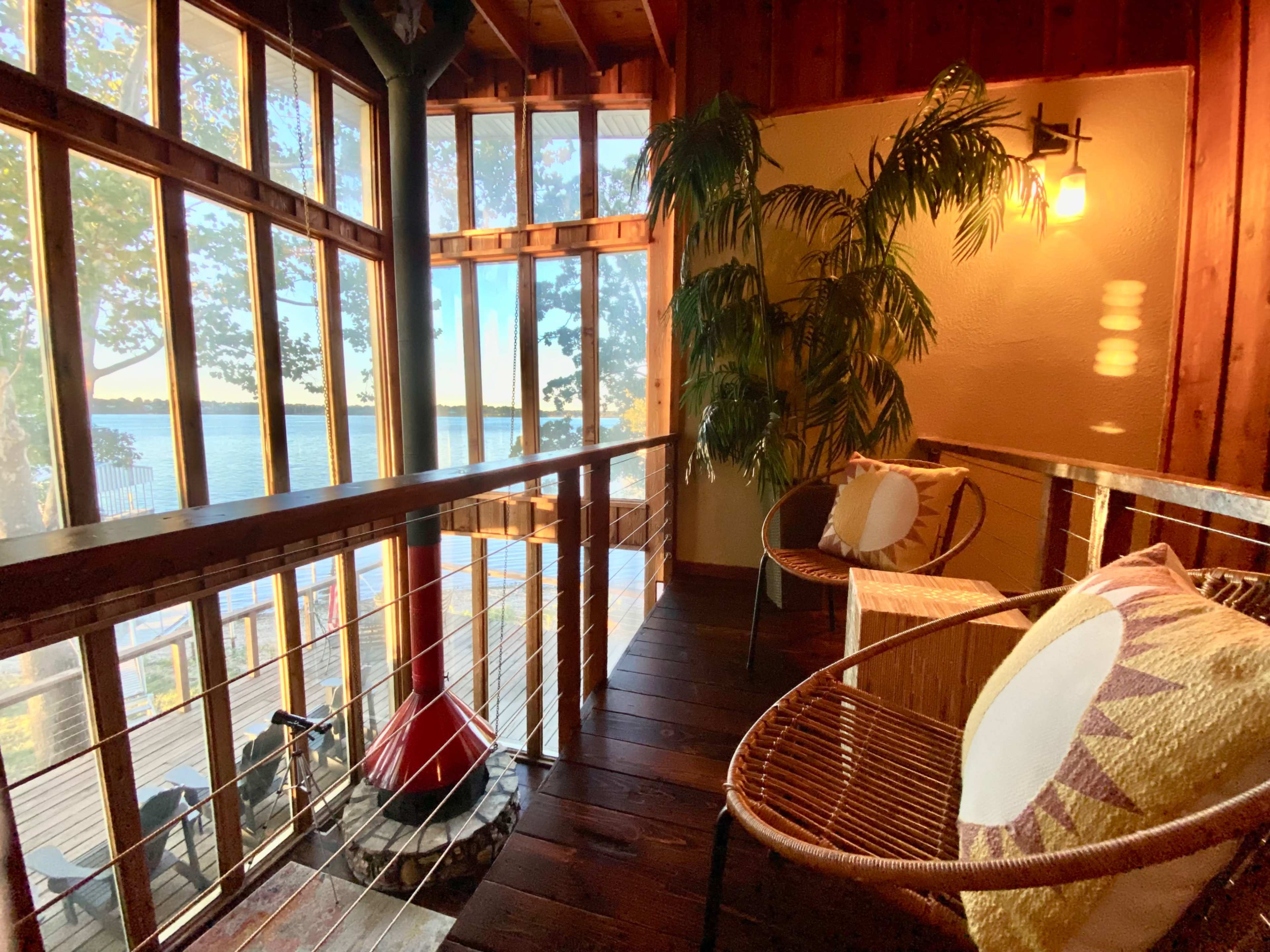 The image shows a cozy interior with large windows overlooking a body of water, featuring a few chairs and a potted plant.