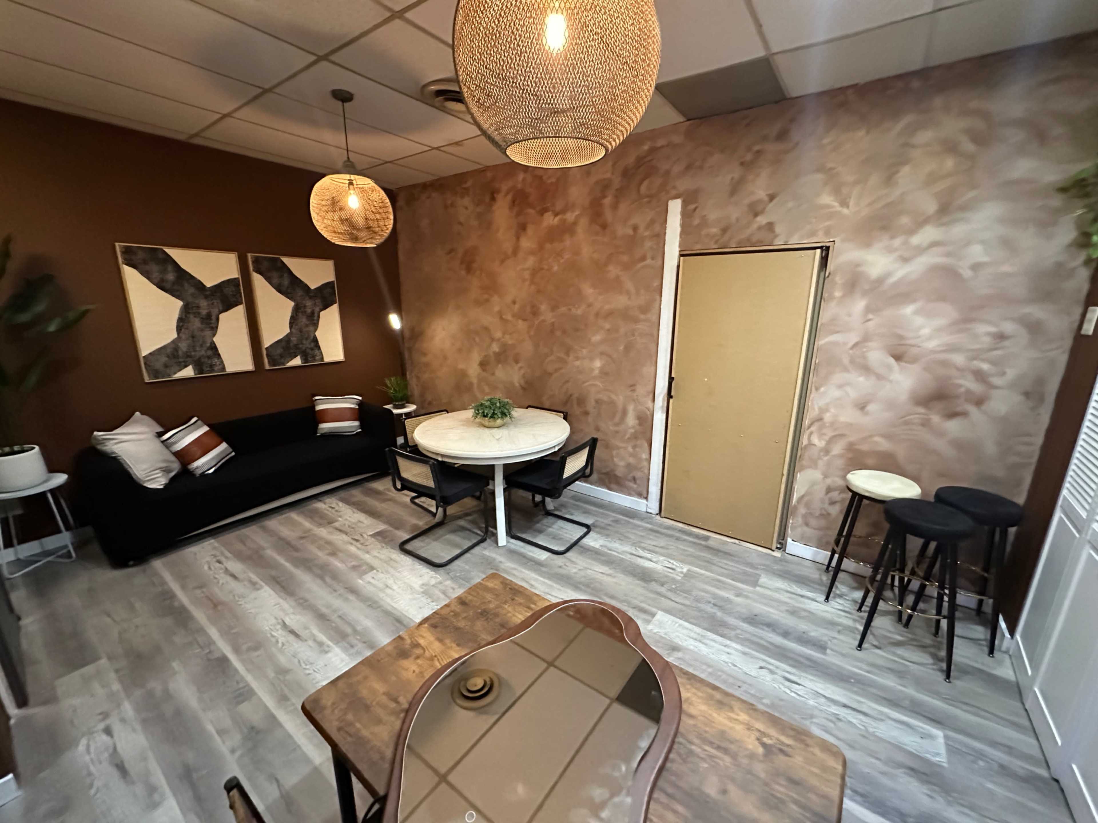 The image shows a modern, well-decorated room featuring a circular dining table, a sofa, two pendant lights, and a textured wall with earthy tones.