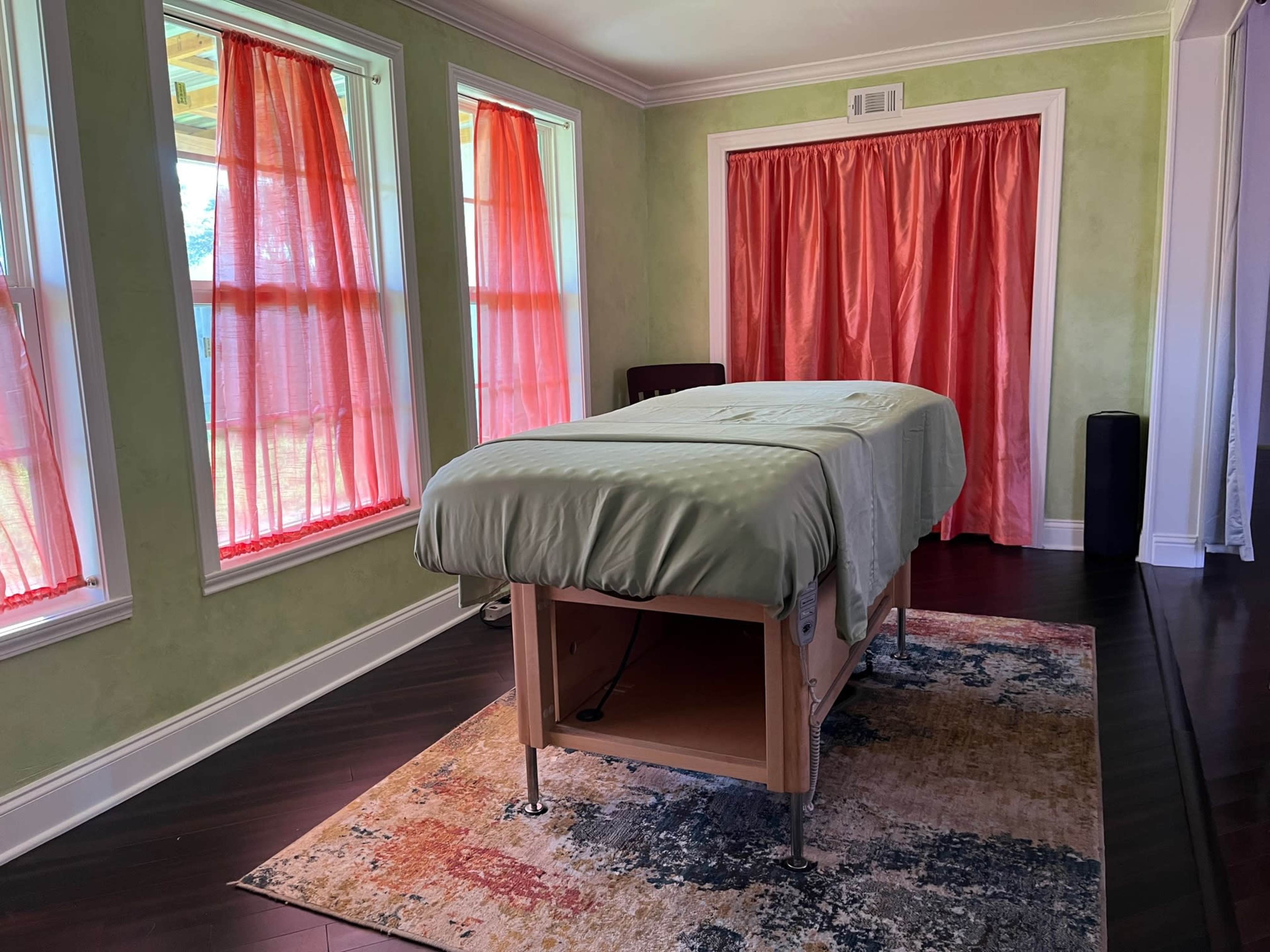 A massage room with a massage table covered in a green sheet, surrounded by windows with red curtains and a patterned rug on the dark wooden floor.