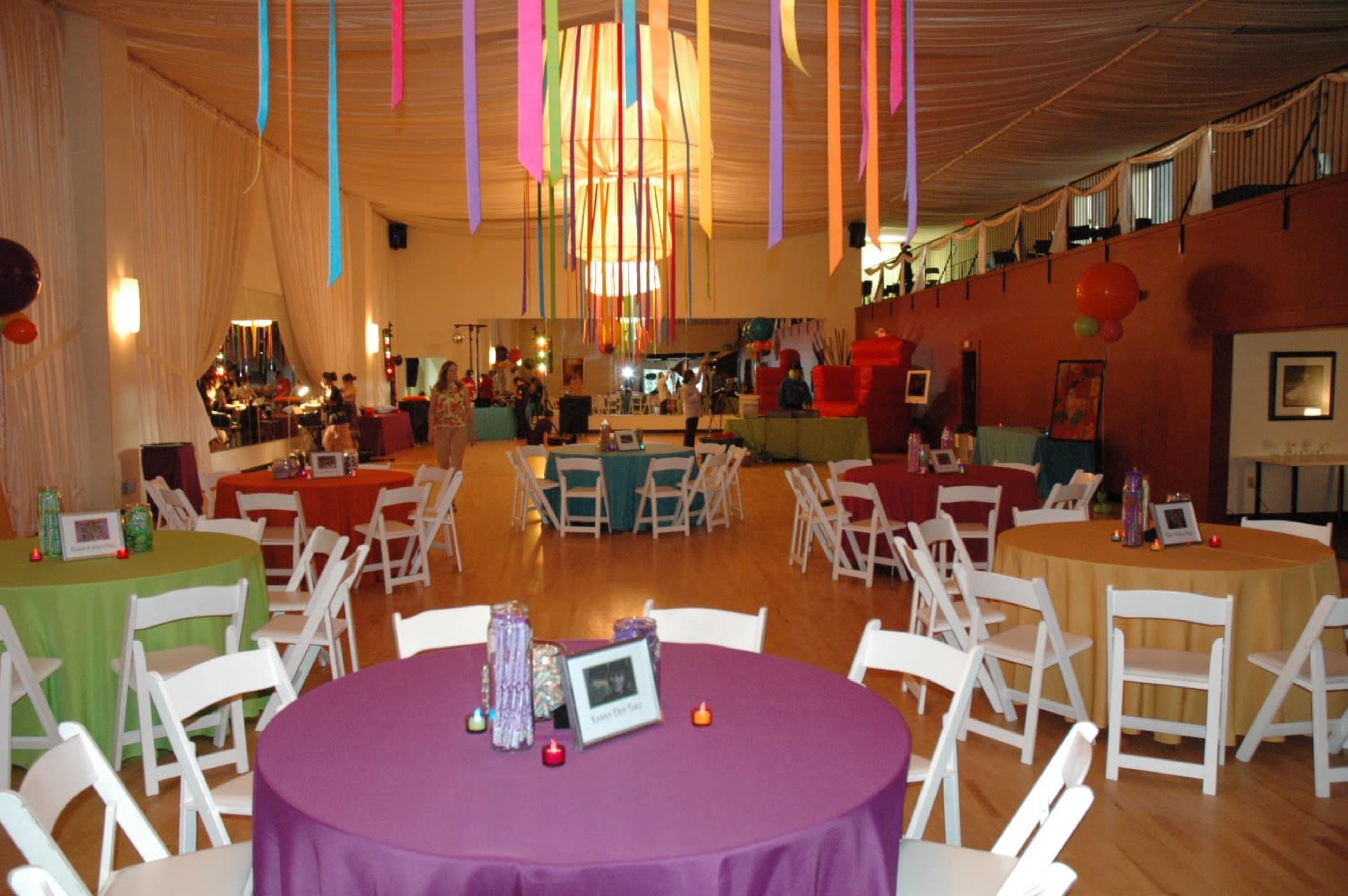 The image shows a brightly decorated event space with round tables covered in various colored tablecloths and streamers hanging from the ceiling.