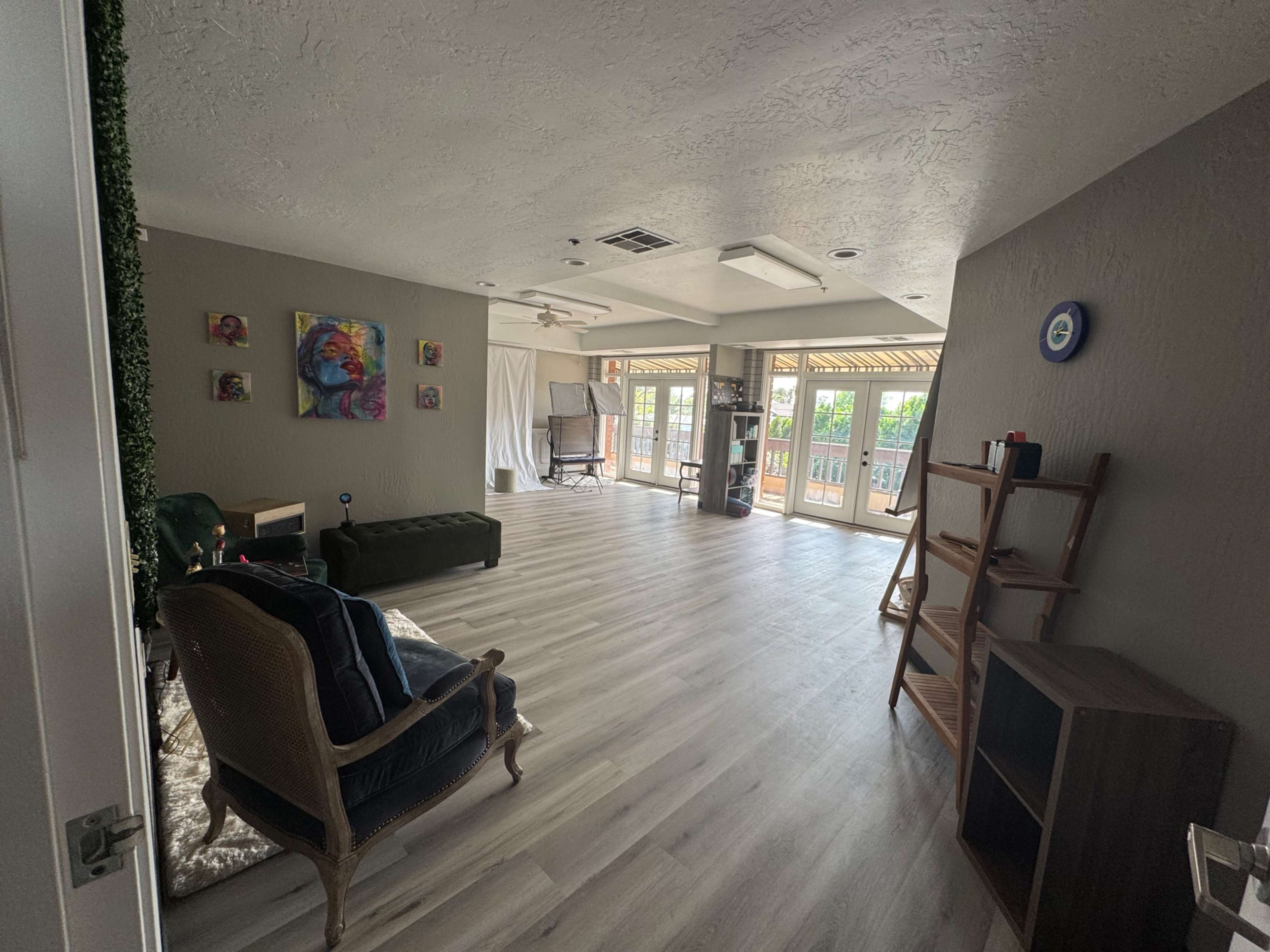 The image depicts a spacious living area with light-colored wood flooring, featuring a green sofa, a chair, and access to a balcony.
