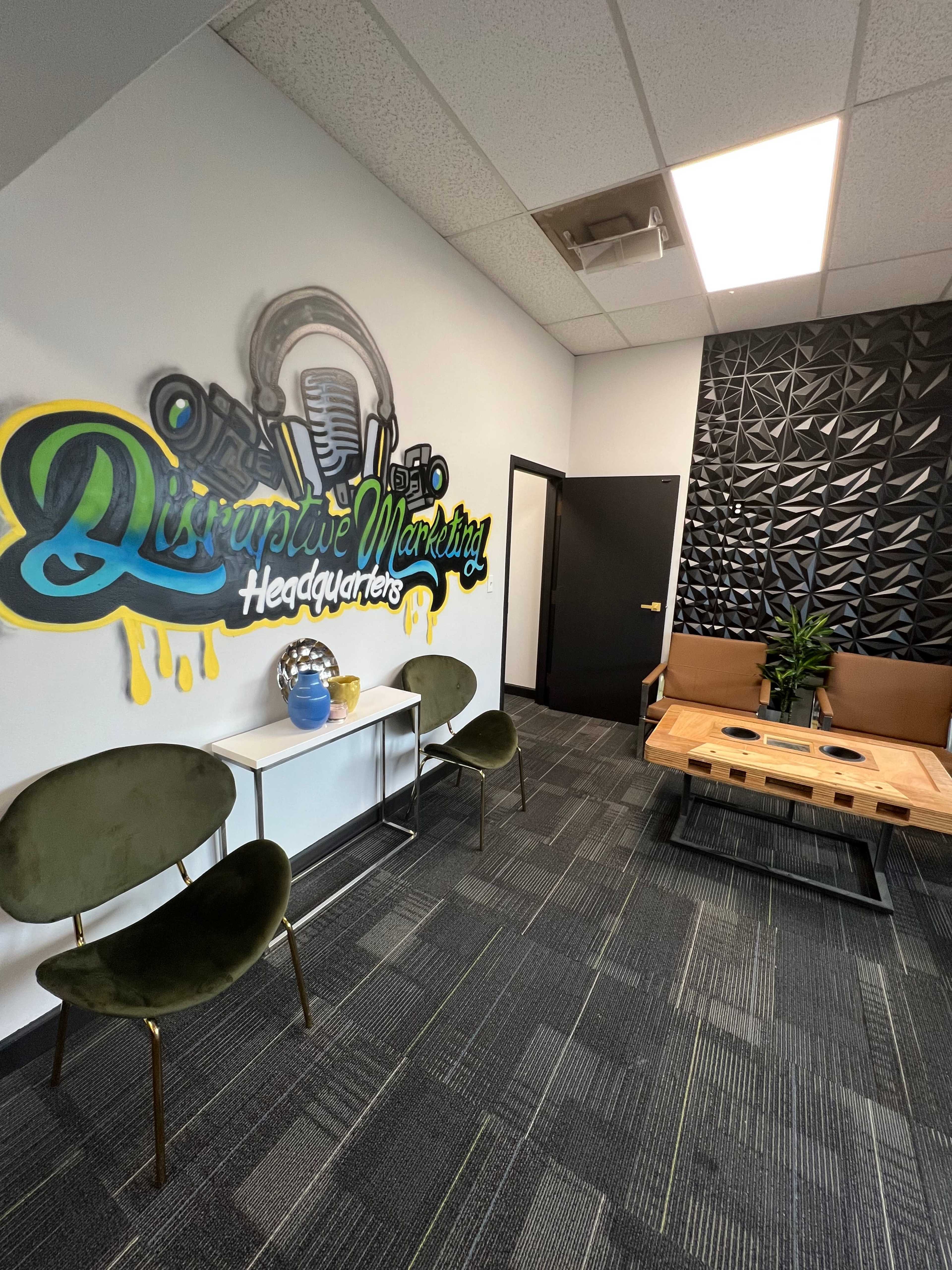 A modern office space with a mural reading "Disruptive Marketing Headquarters," green chairs, a wooden coffee table, and a decorative plant.