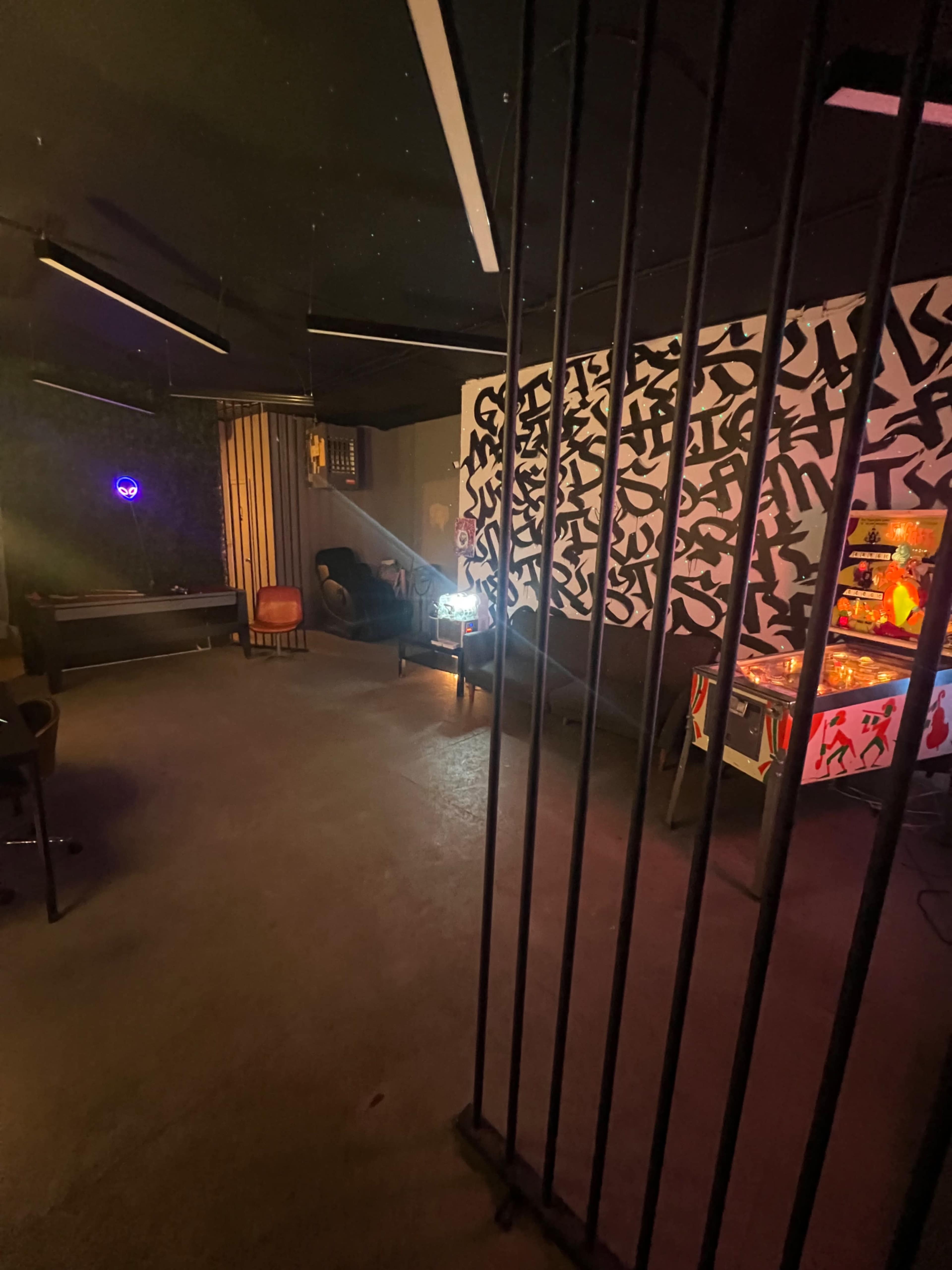 The image shows a dimly lit room with a pinball machine, contemporary furniture, and a patterned wall behind bars.