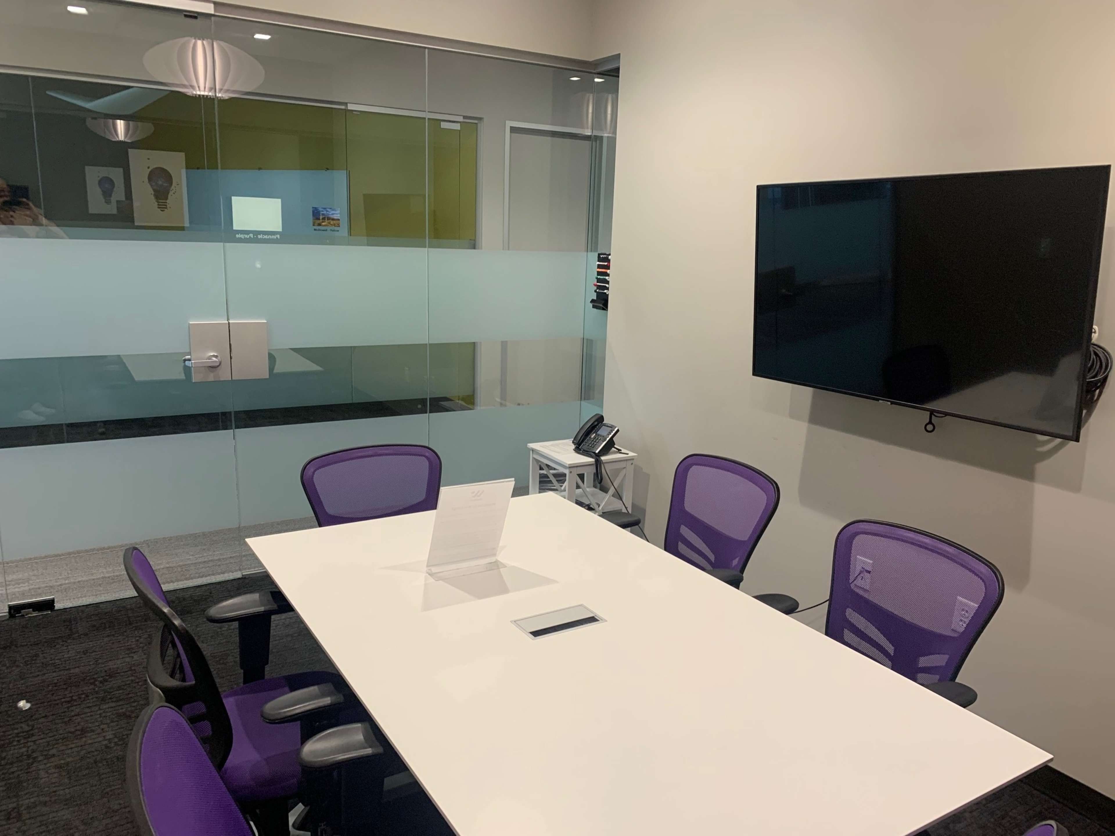 A meeting room features a white table surrounded by purple chairs and a mounted television on the wall.