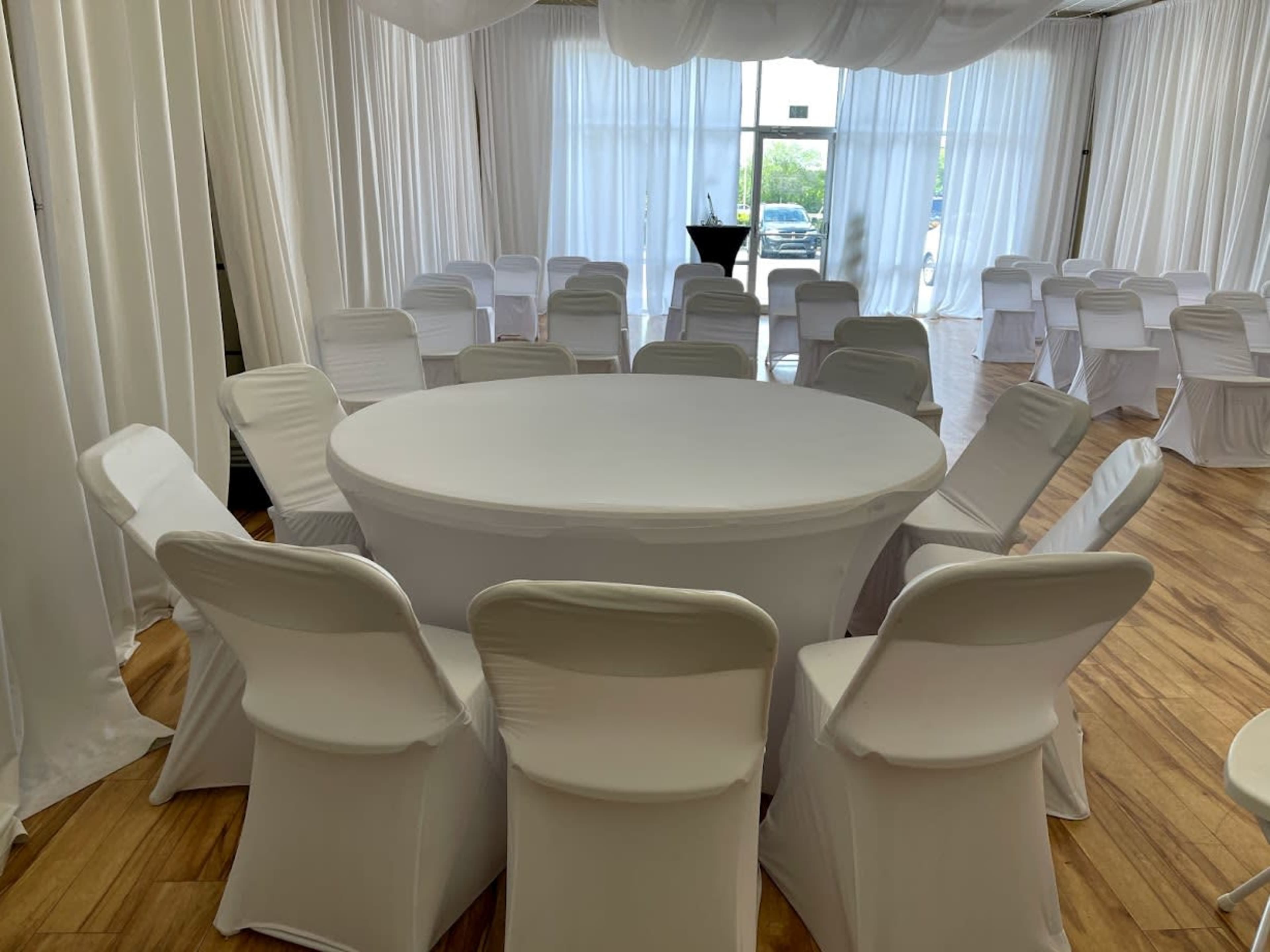 A round table covered with a white tablecloth is surrounded by a set of white chairs in a softly lit indoor space with draped white fabric.