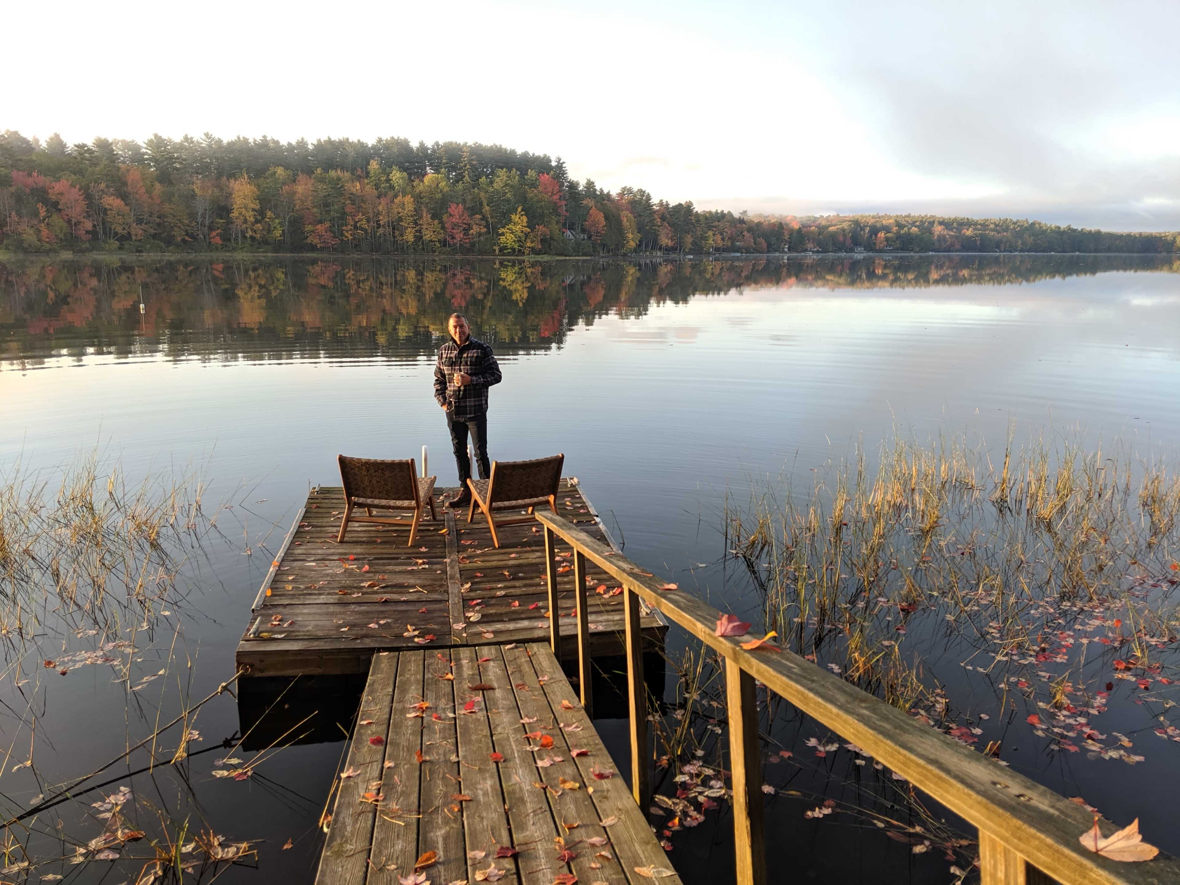 A man stands on a wooden dock surrounded by fallen leaves, overlooking a calm lake reflecting autumn trees in the distance.