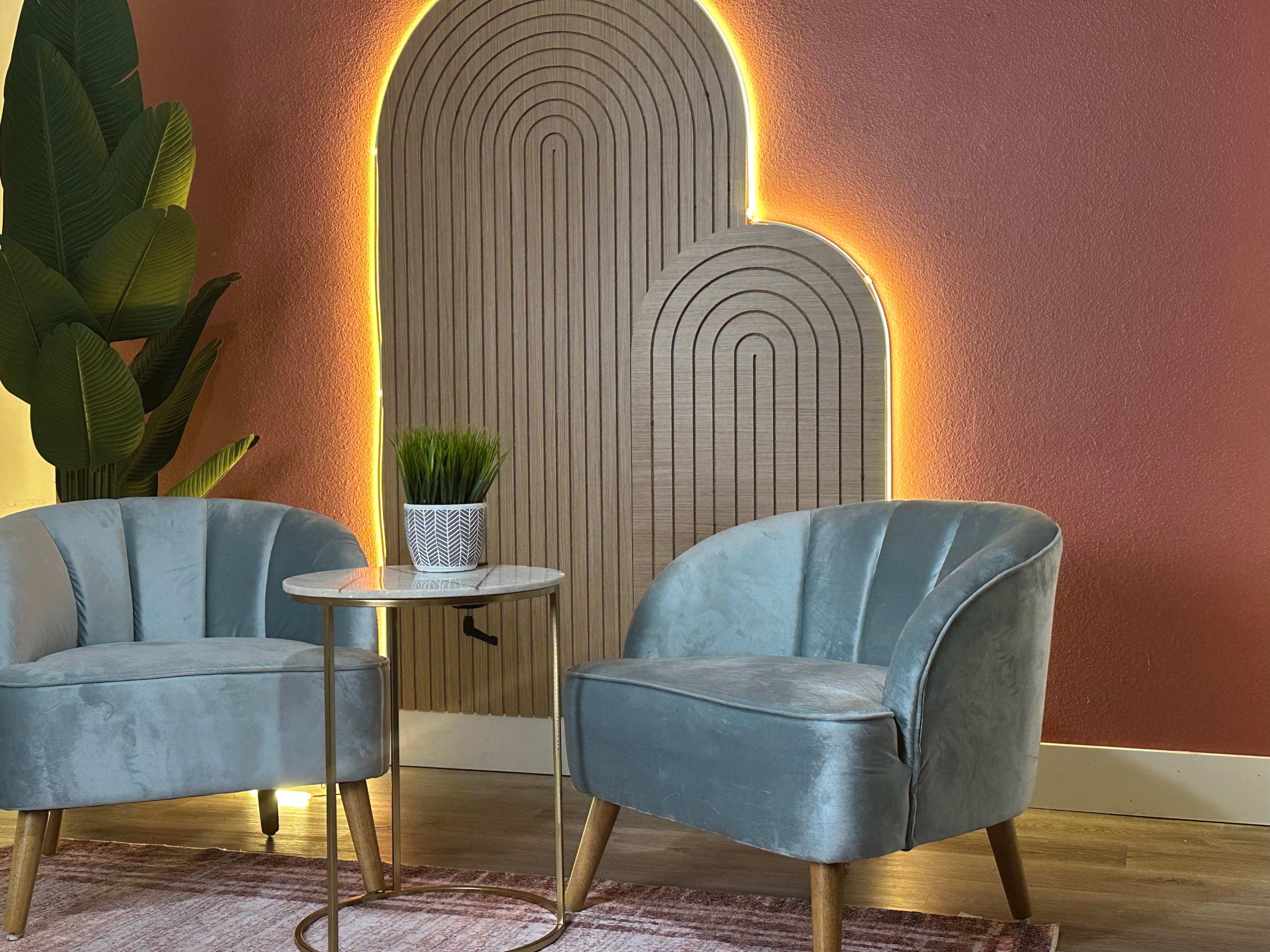 The image shows two velvet armchairs positioned beside a small round table, with a decorative wall featuring a glowing, layered wooden design and a potted plant nearby.