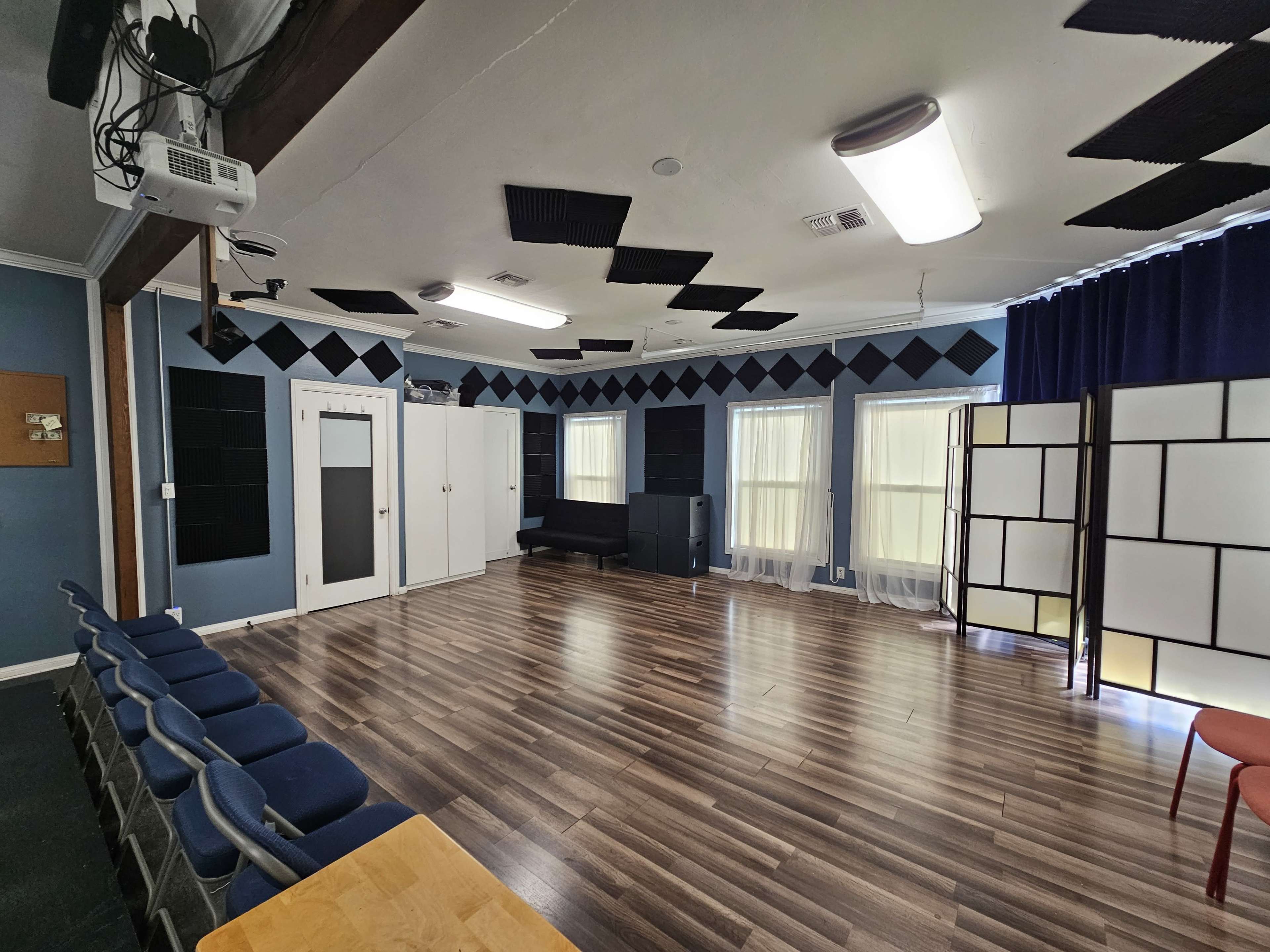 The image shows a spacious room with wooden flooring, acoustic panels, a piano, and seating arranged in rows.