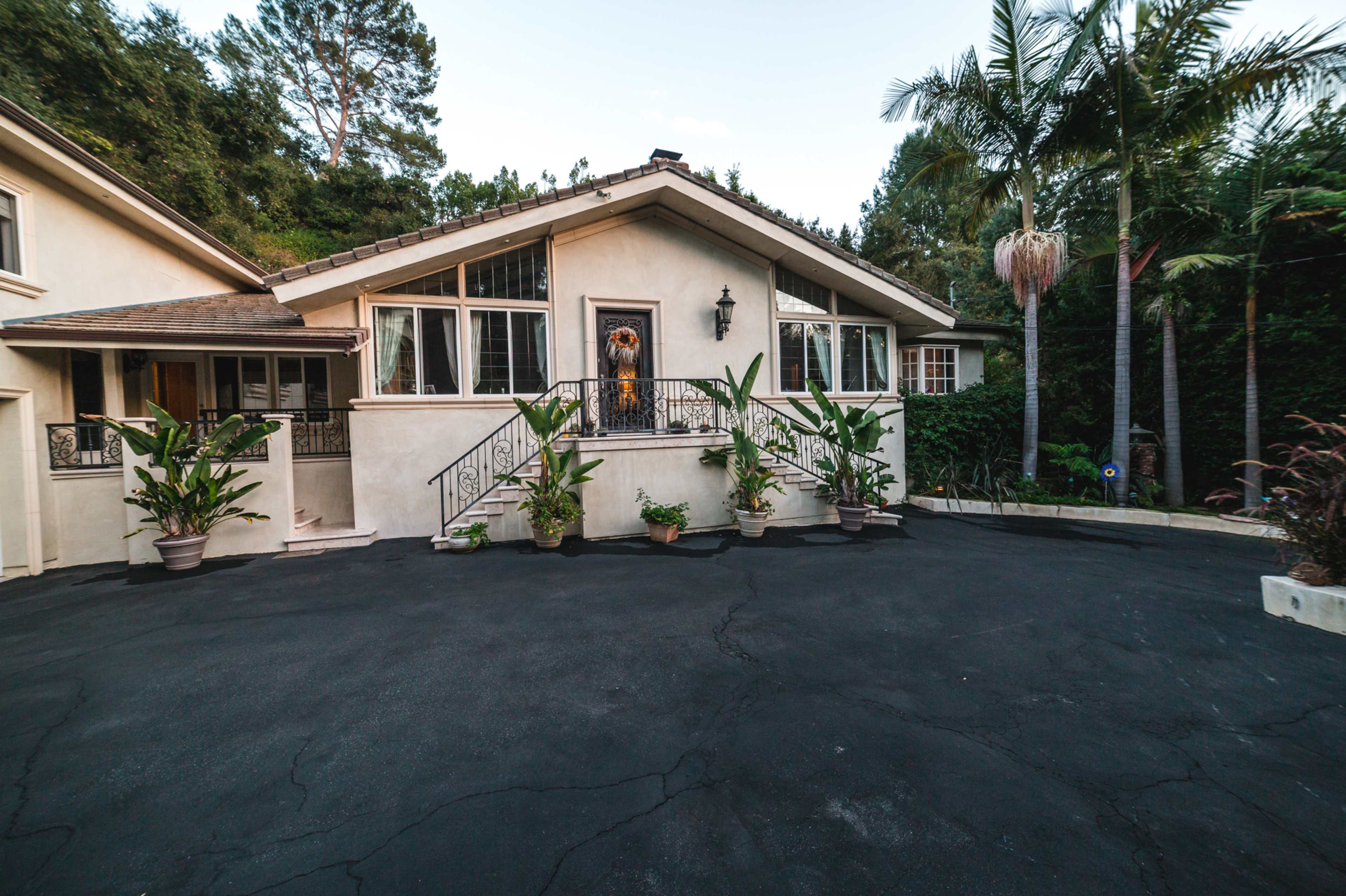 A house with a front porch and large windows is surrounded by potted plants and palm trees on a black asphalt driveway.