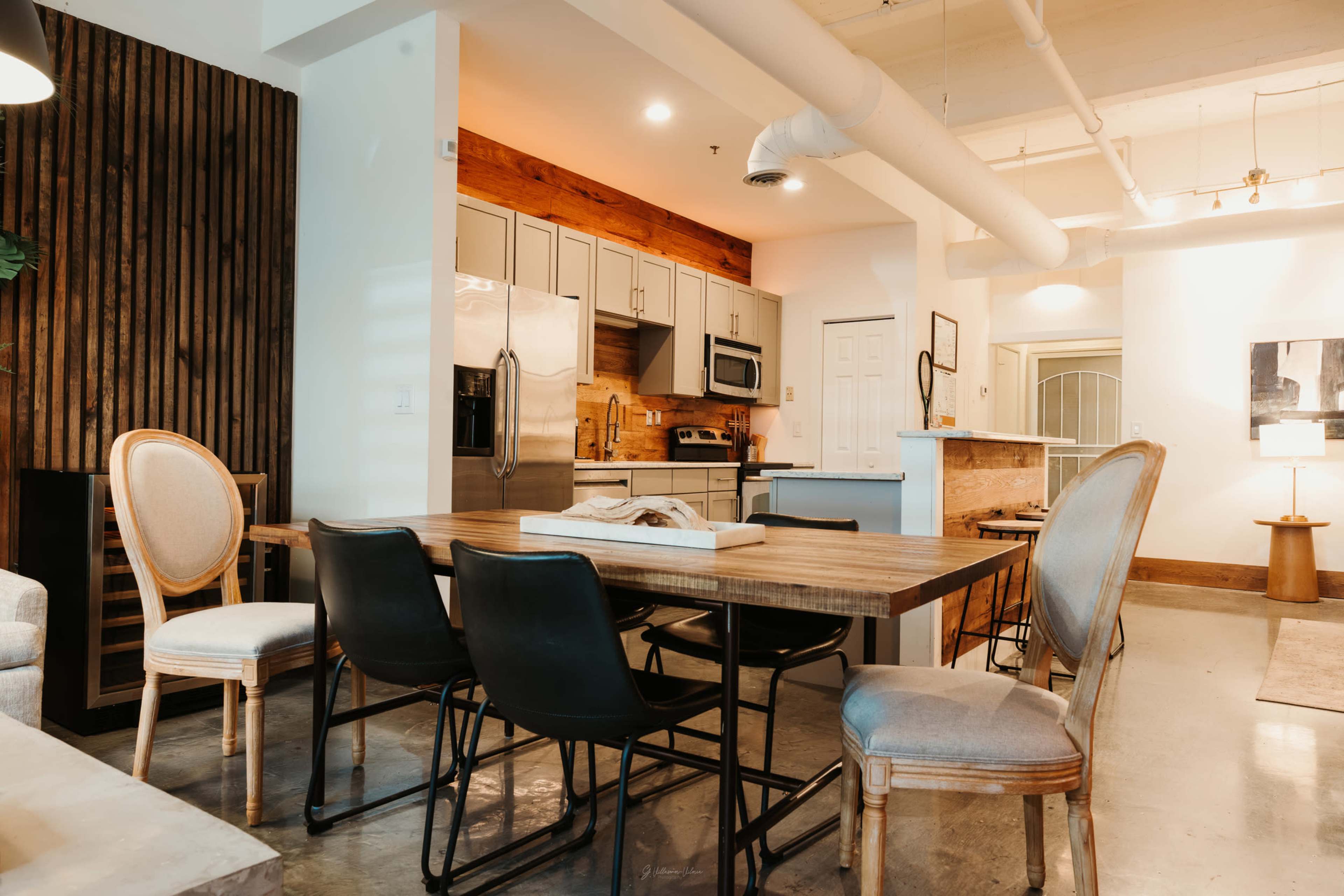 The image shows a modern dining area featuring a wooden table surrounded by black and light-colored chairs, with an open kitchen in the background.