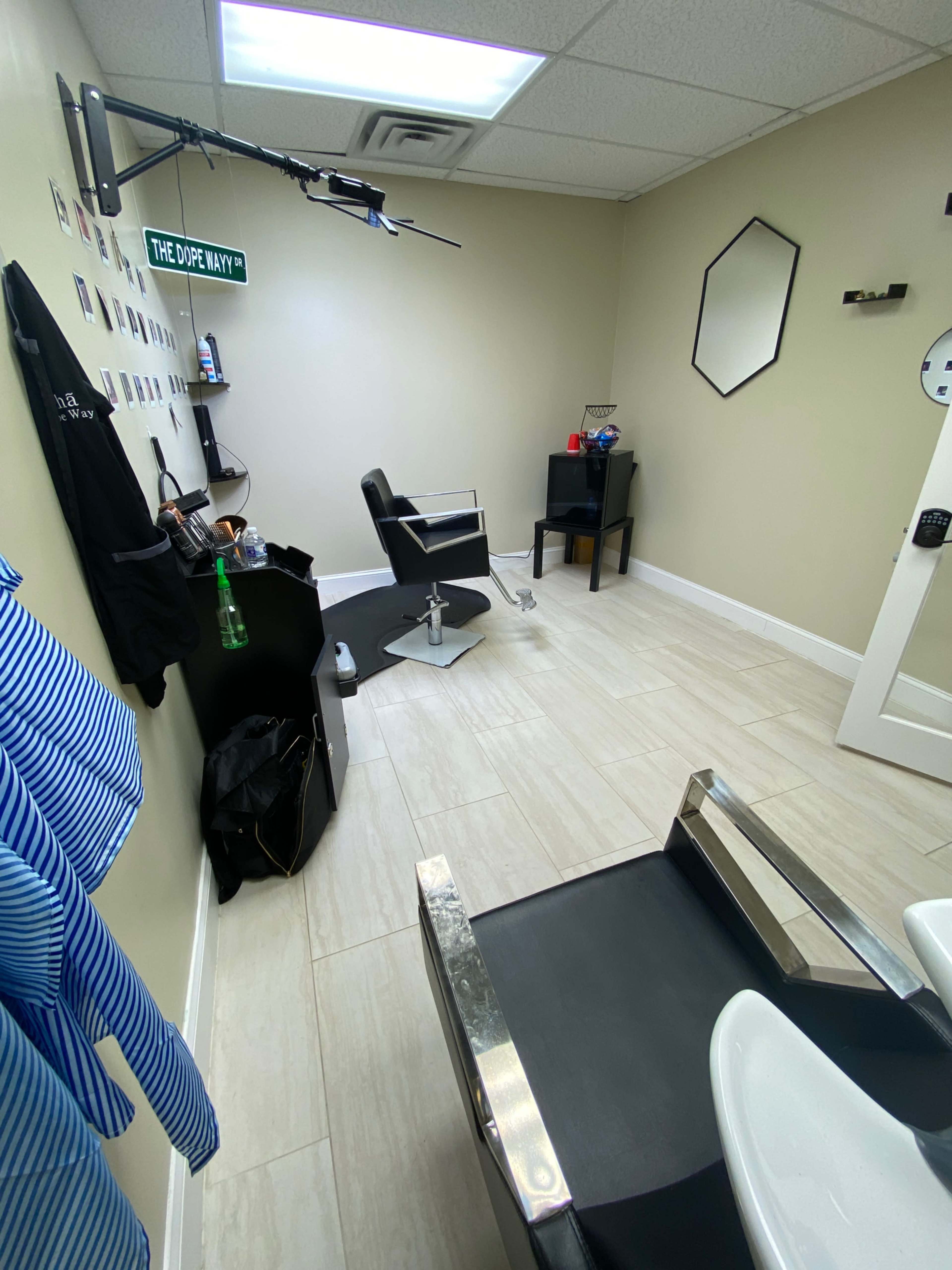 The image shows a small hair salon interior featuring a black barber chair, a styling station with a mirror, and a neatly arranged wall with photographs and a sign reading "THE DOPE WAY."