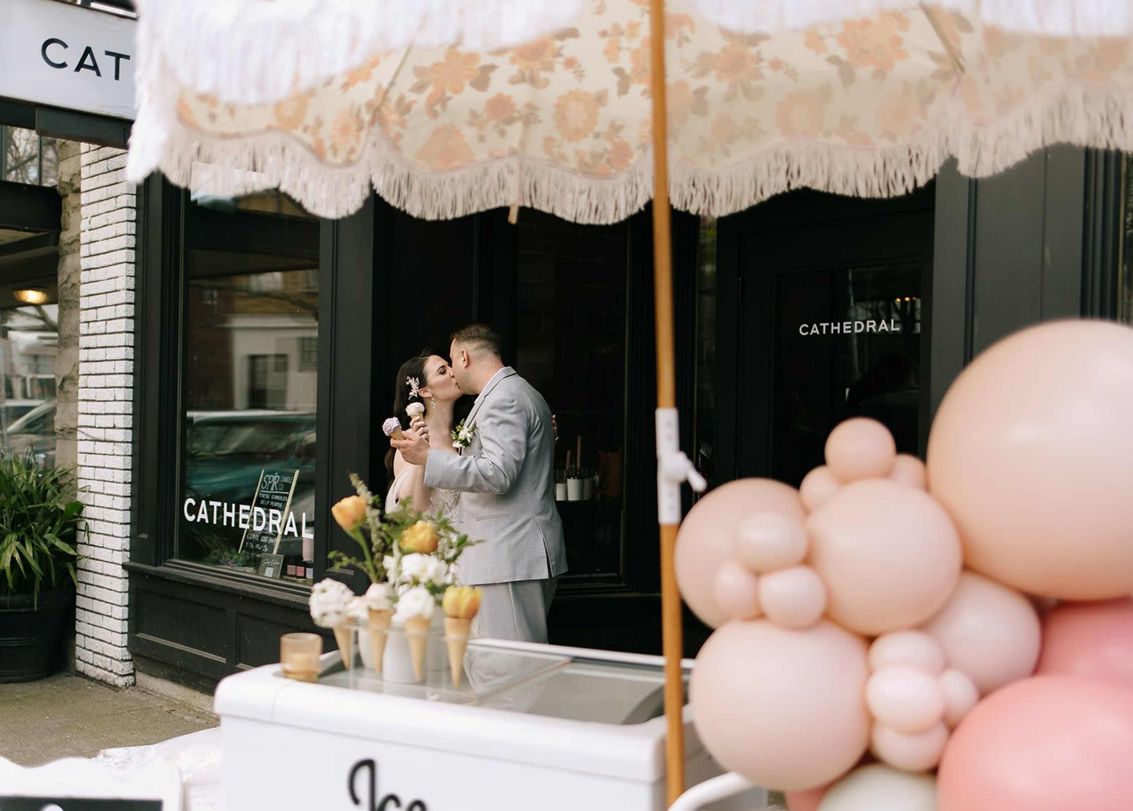 A couple kisses under a decorative umbrella near an ice cream cart adorned with balloons outside a storefront.