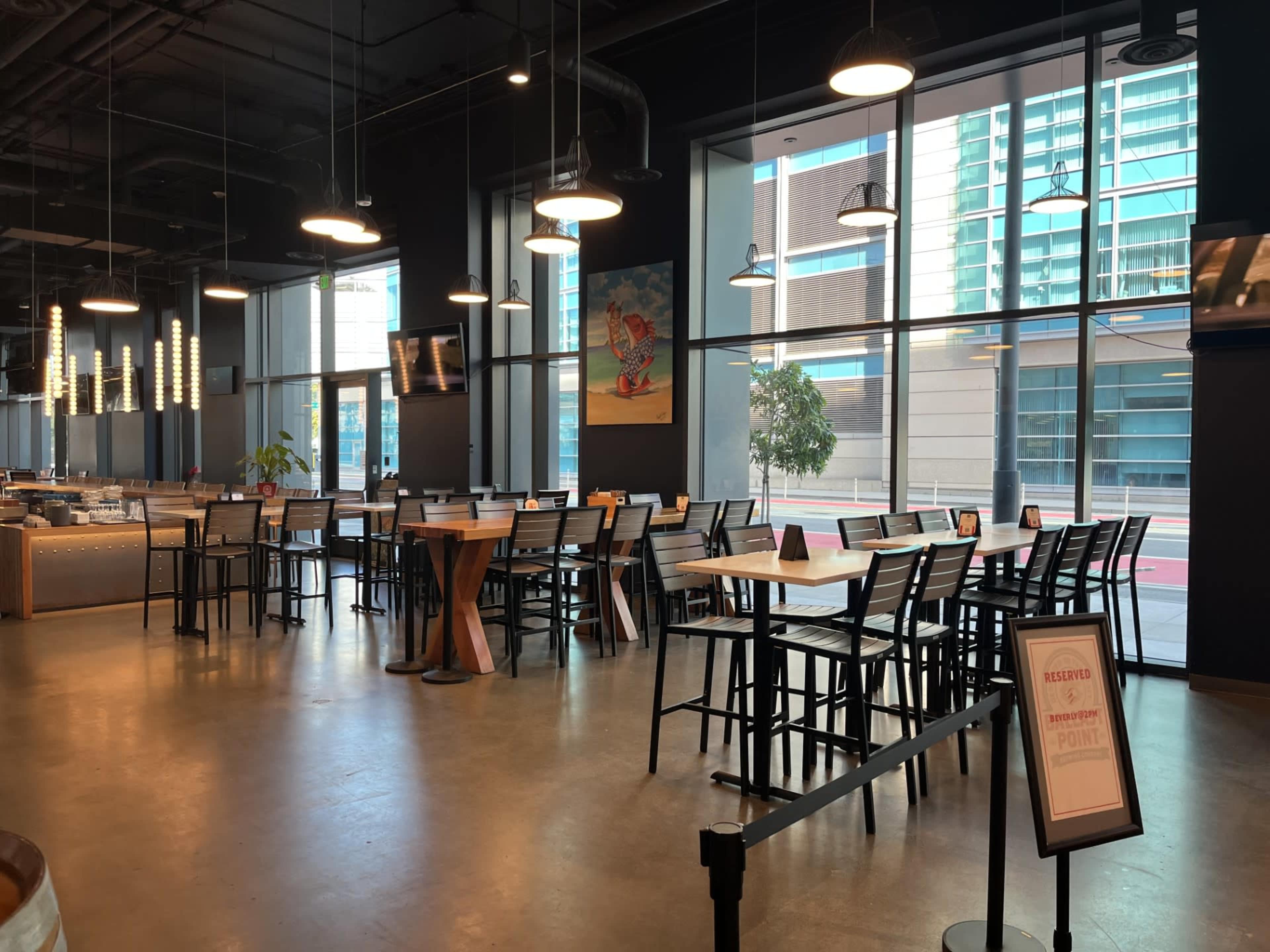 The image shows a spacious indoor dining area with tall tables and chairs, large windows providing natural light, and a few decorative elements on the walls.