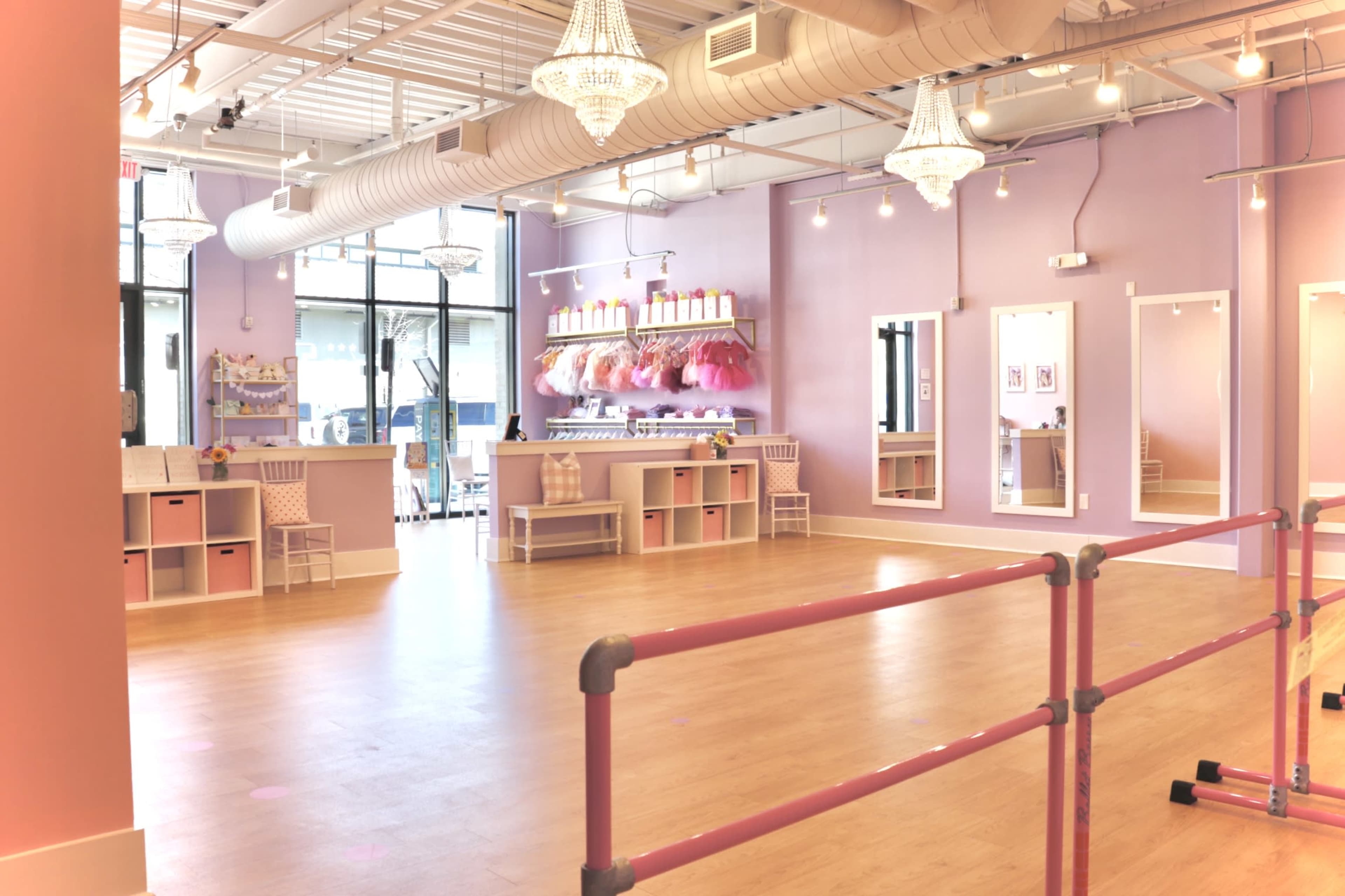 The image shows a bright dance studio with pink walls, a wooden floor, mirrored walls, and a barre in the center, featuring a reception area with shelves and colorful costumes.