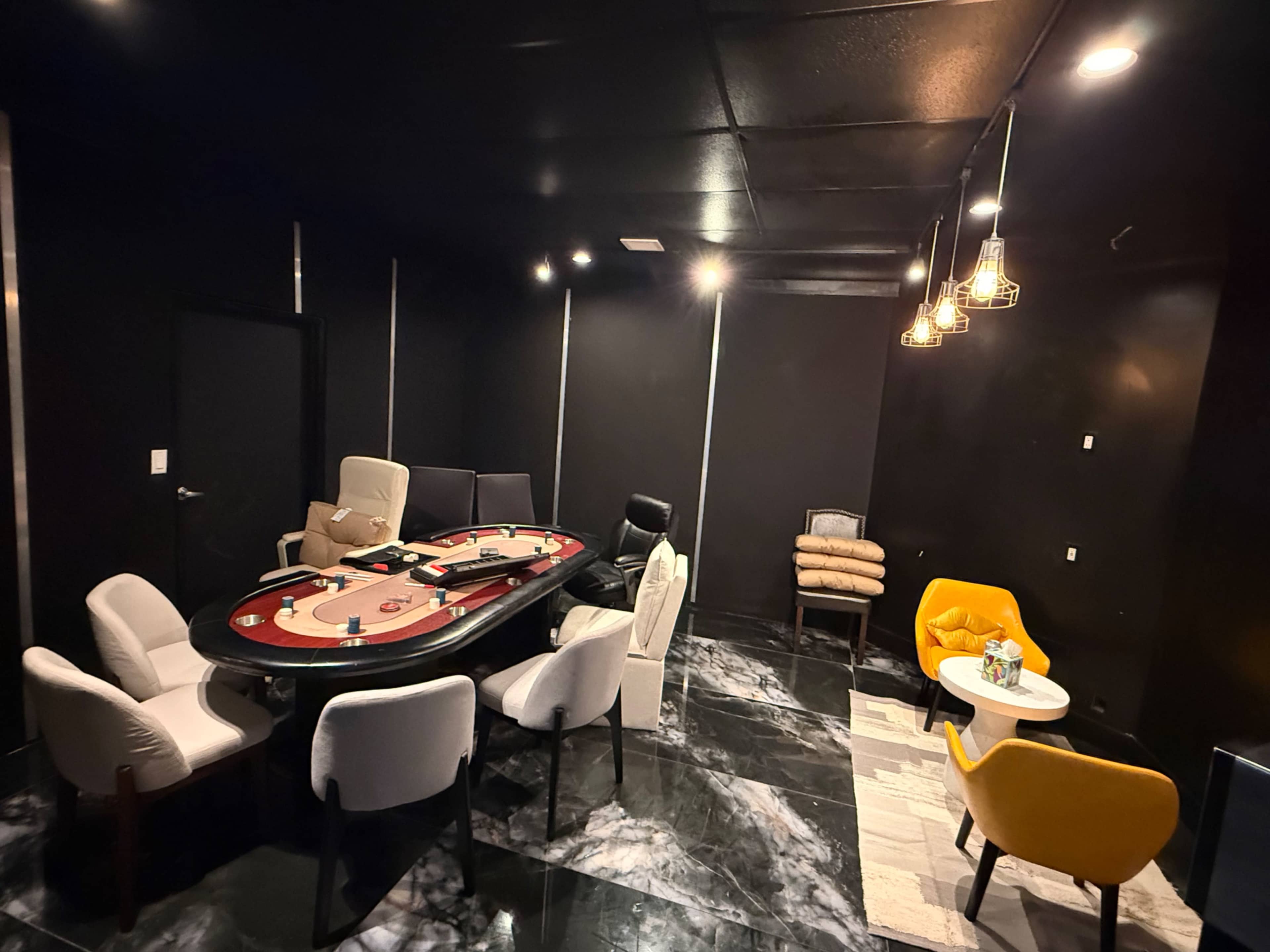 The image shows a dimly lit room featuring a round poker table surrounded by several chairs, with a small seating area in the corner that includes a yellow chair and a side table.