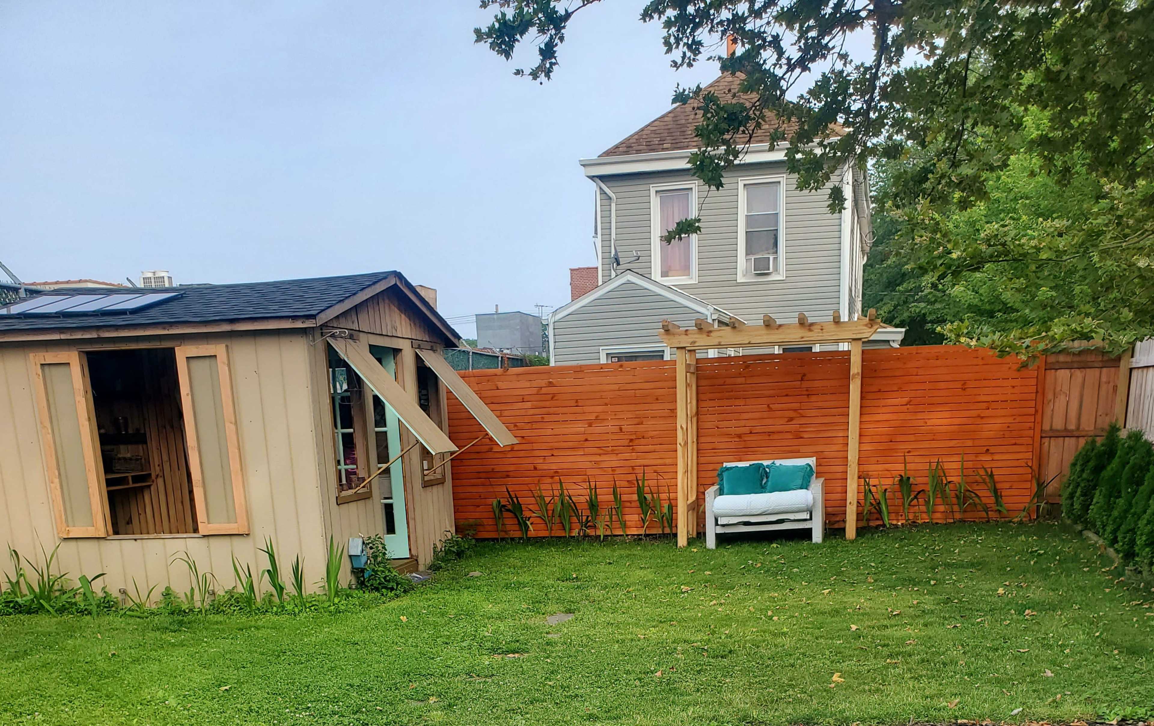 The image shows a backyard scene featuring a small shed with an open window, a wooden fence, a white bench with cushions, and a house in the background.