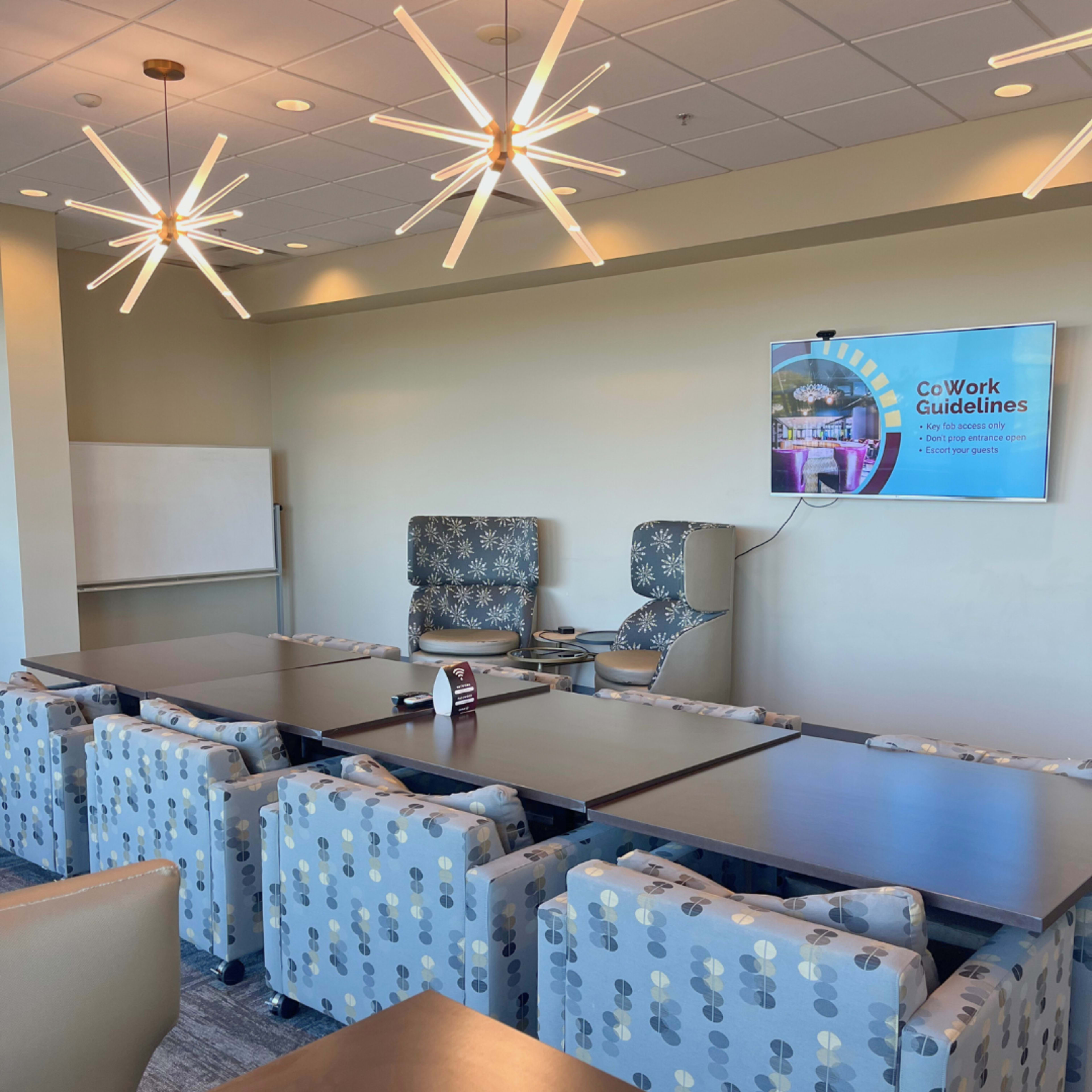 The image shows a modern conference room with a long table surrounded by patterned chairs, two stylish armchairs, and a screen displaying "CoWork Guidelines" on the wall.