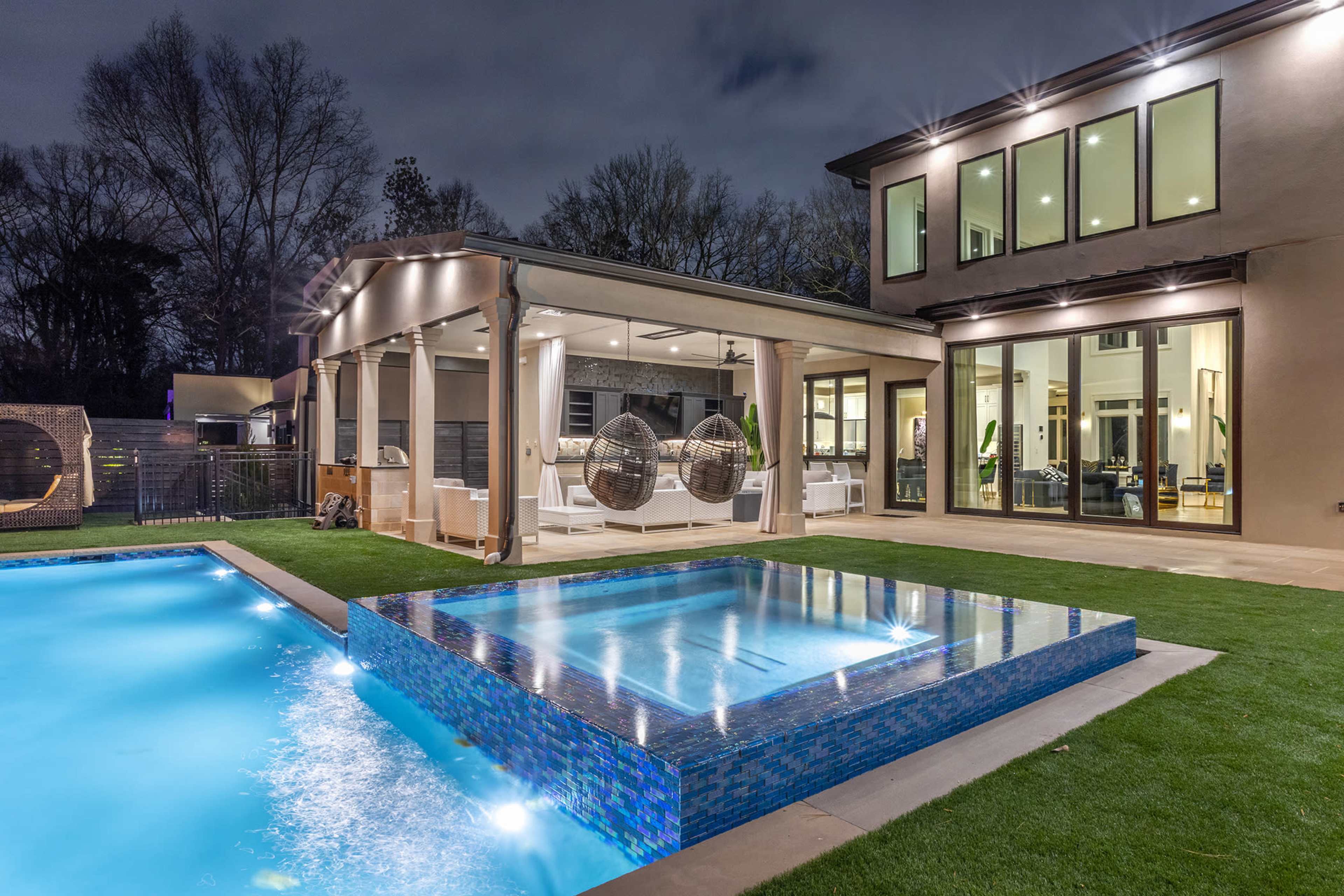 A modern backyard with a swimming pool and a hot tub, surrounded by manicured grass and a patio with overhead seating.