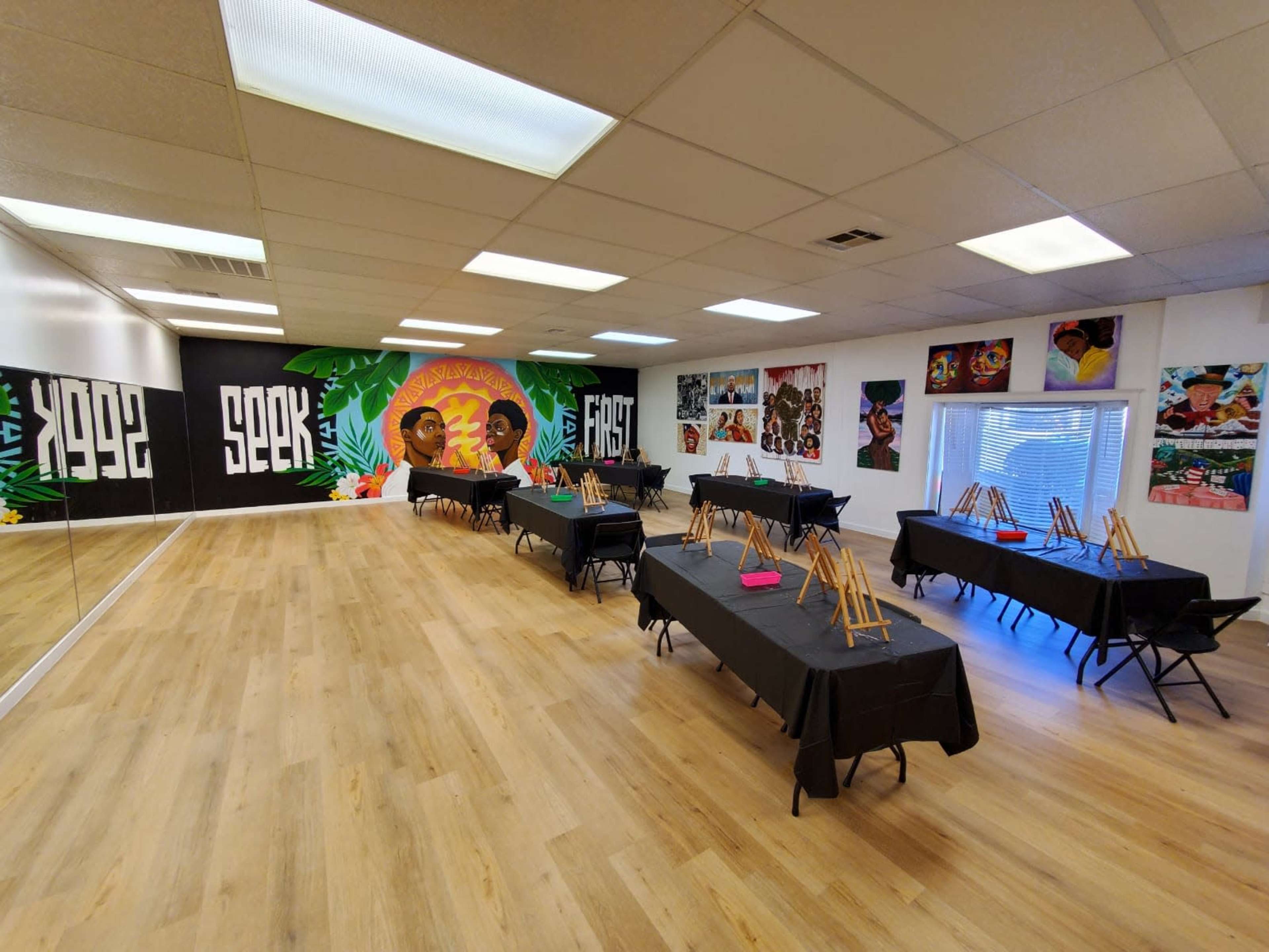 The image shows an art studio with several black tables set up for an event, each equipped with easels, surrounded by colorful murals and artwork on the walls.