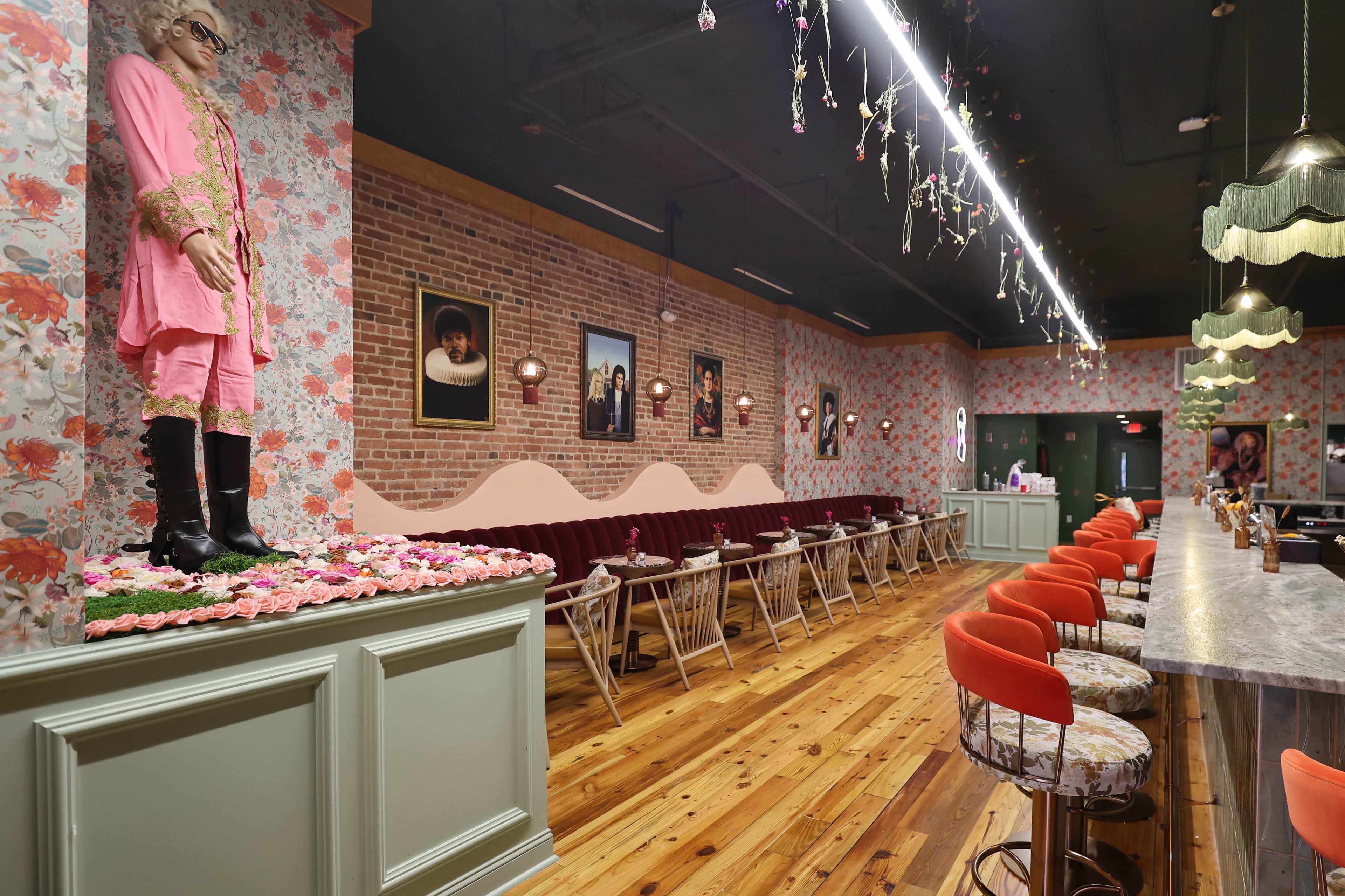 The image shows a stylish cafe with pink decor, wooden flooring, and a mannequin dressed in pink attire displayed on a floral-covered counter.