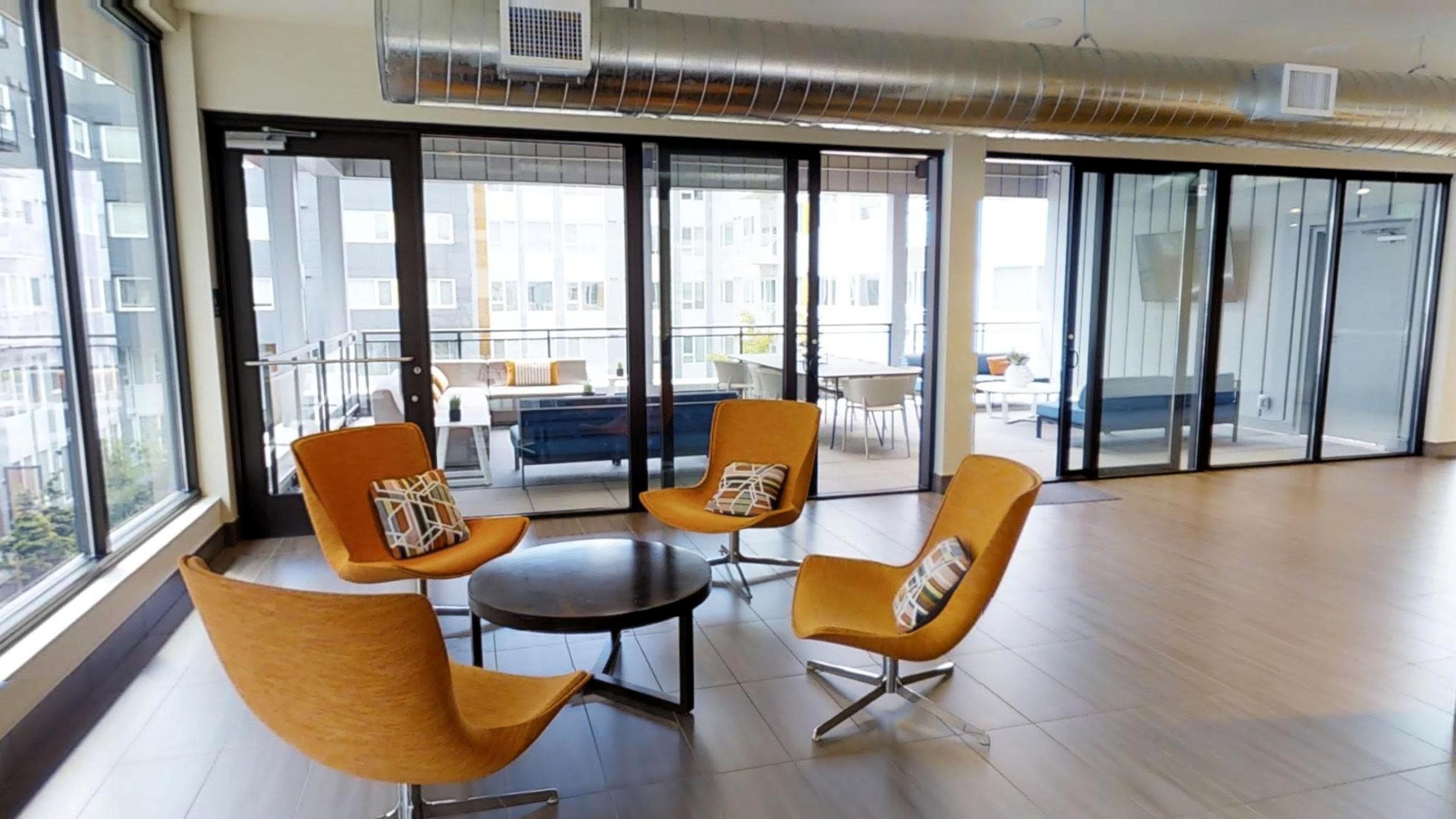 The image shows a modern room with large windows, featuring four orange chairs arranged around a round table.