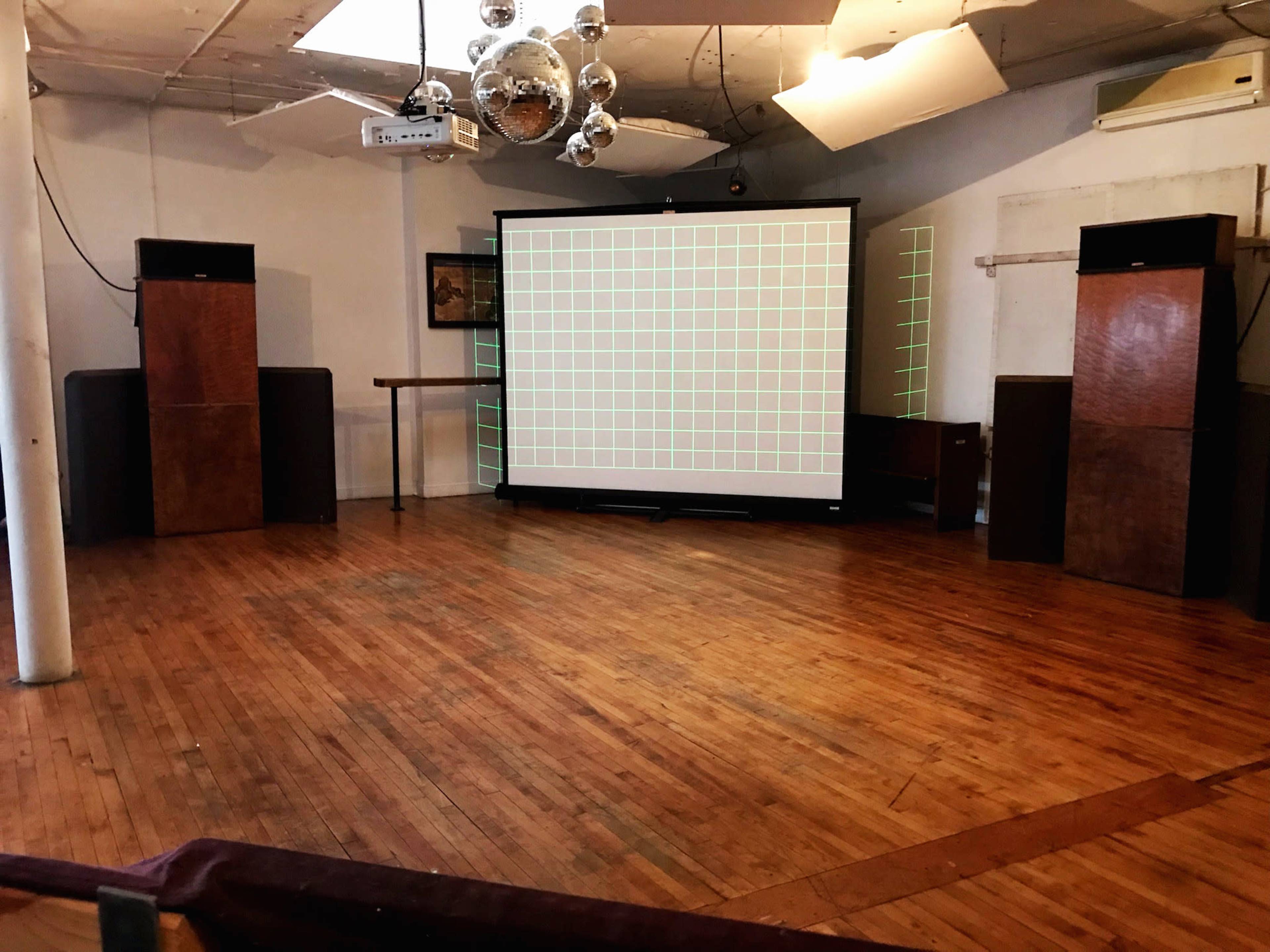 The image shows an empty room with wooden flooring, featuring large speakers on either side and a projection screen at the front displaying a grid pattern.