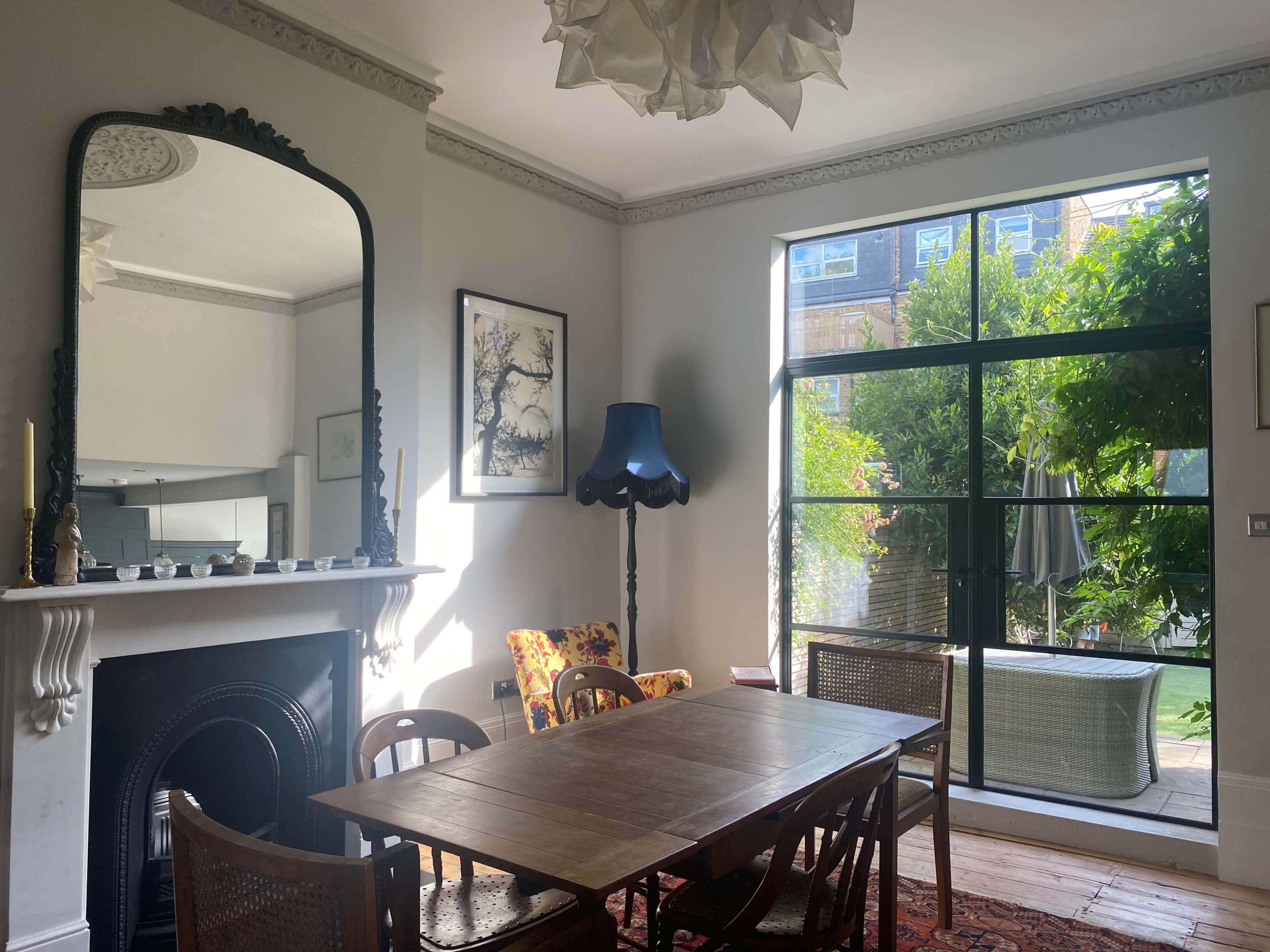 A dining area features a wooden table, chairs, a large mirror, and a window with a view of a garden.