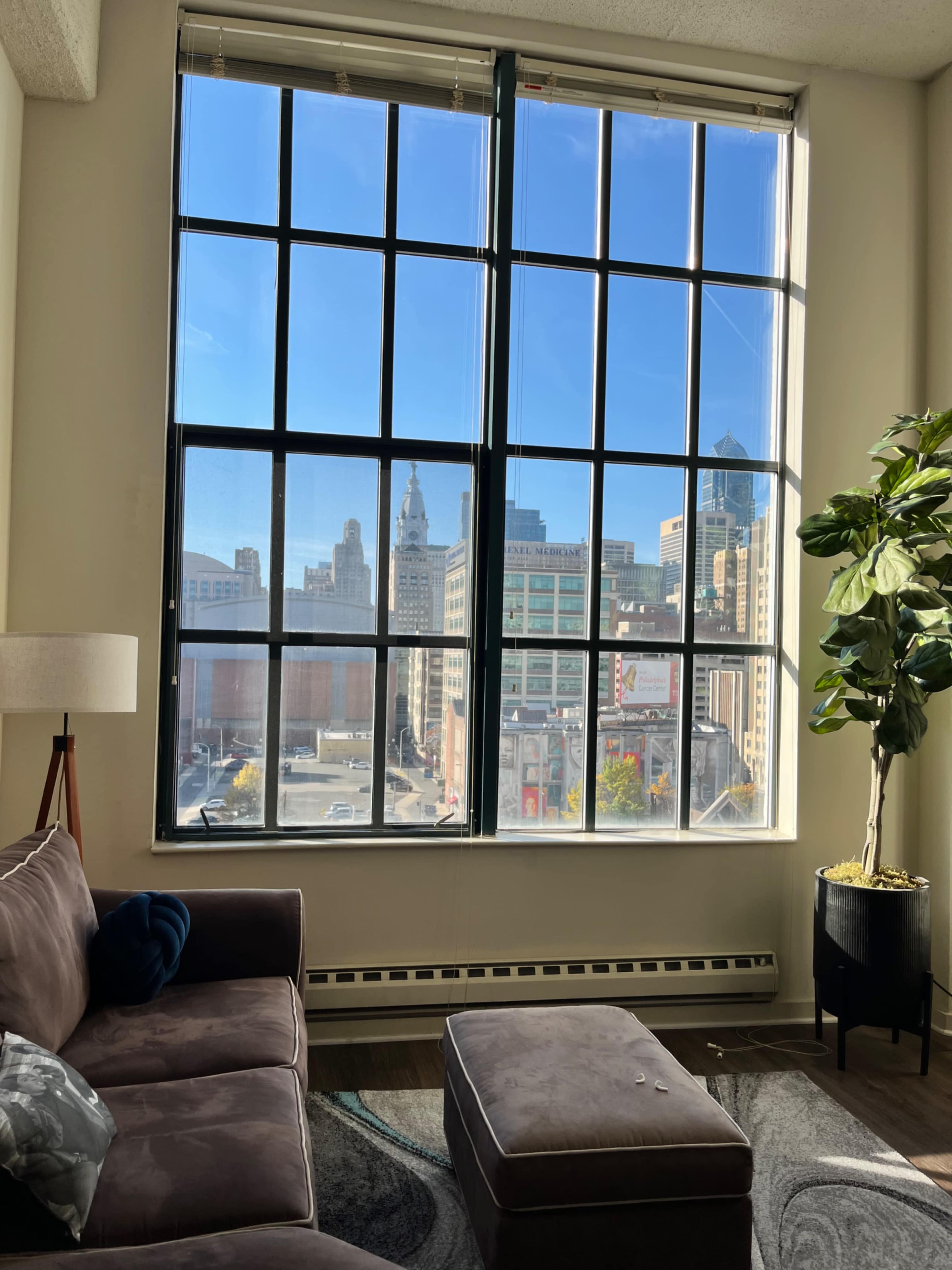The image shows a living room with a large window overlooking a cityscape, featuring buildings under a clear blue sky.