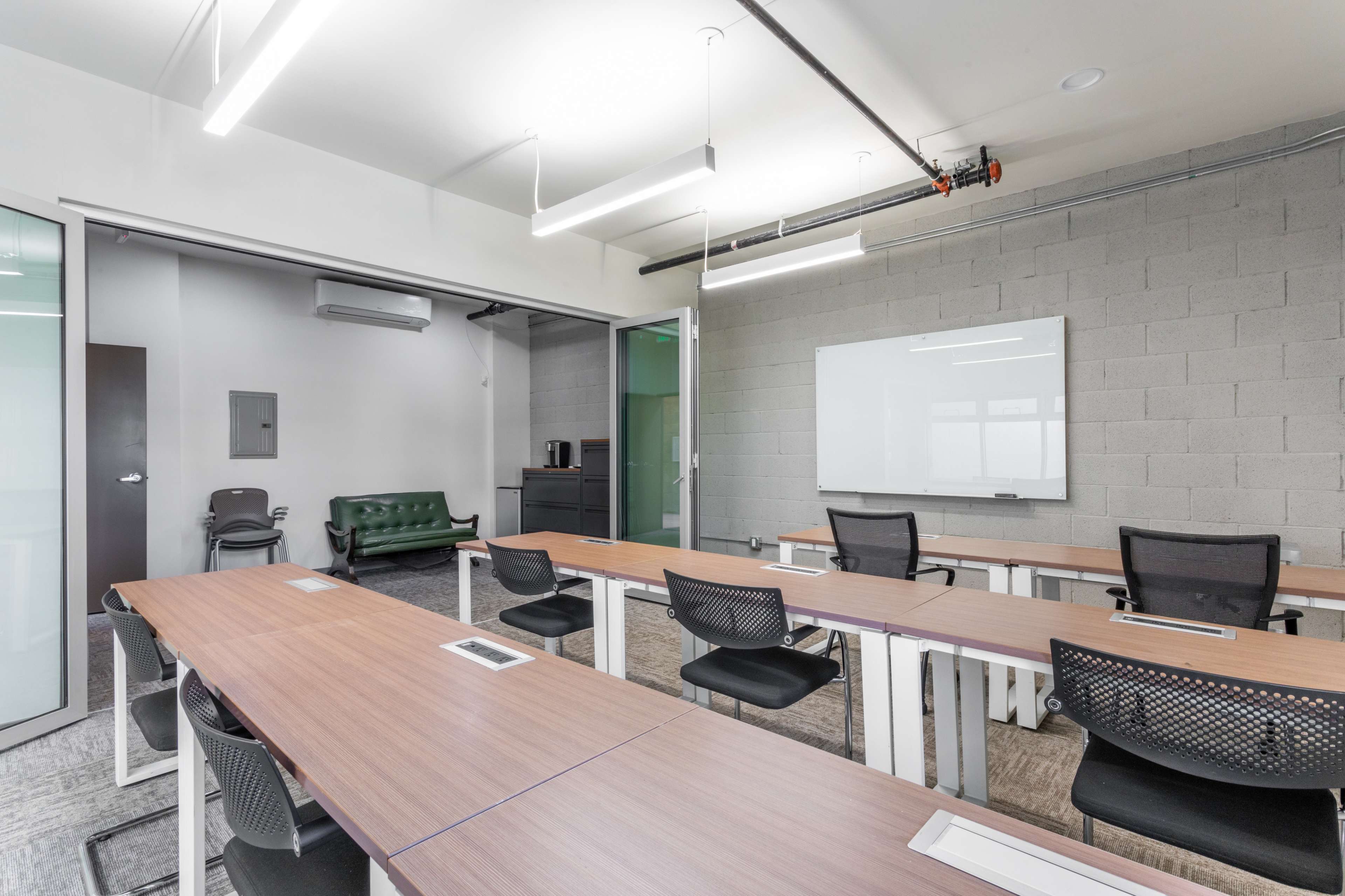 A modern classroom with multiple tables, black chairs, a green couch in an adjoining room, and a whiteboard on the wall.