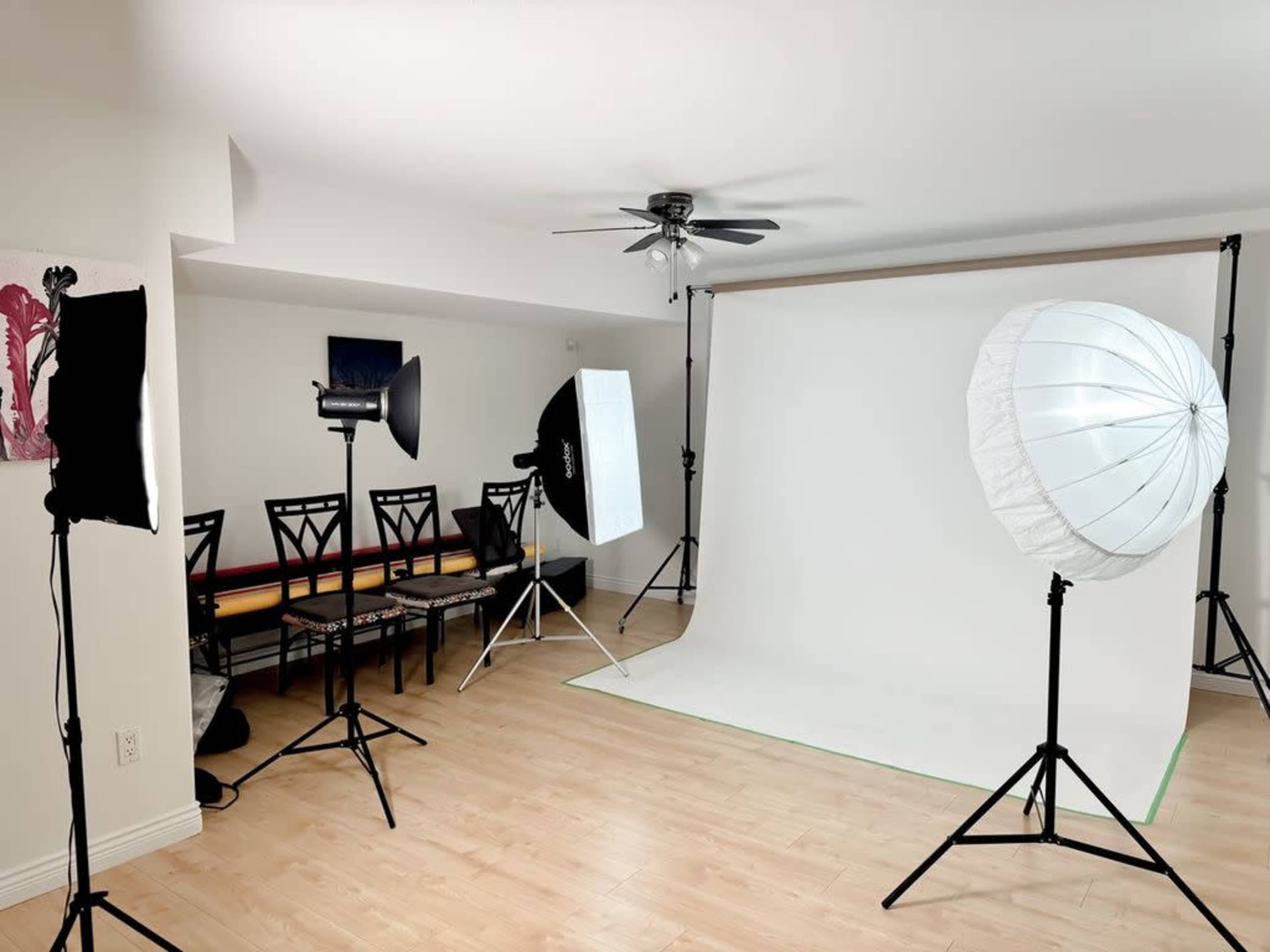 A photography studio is set up with various lights and a white backdrop, along with a row of chairs against the wall.