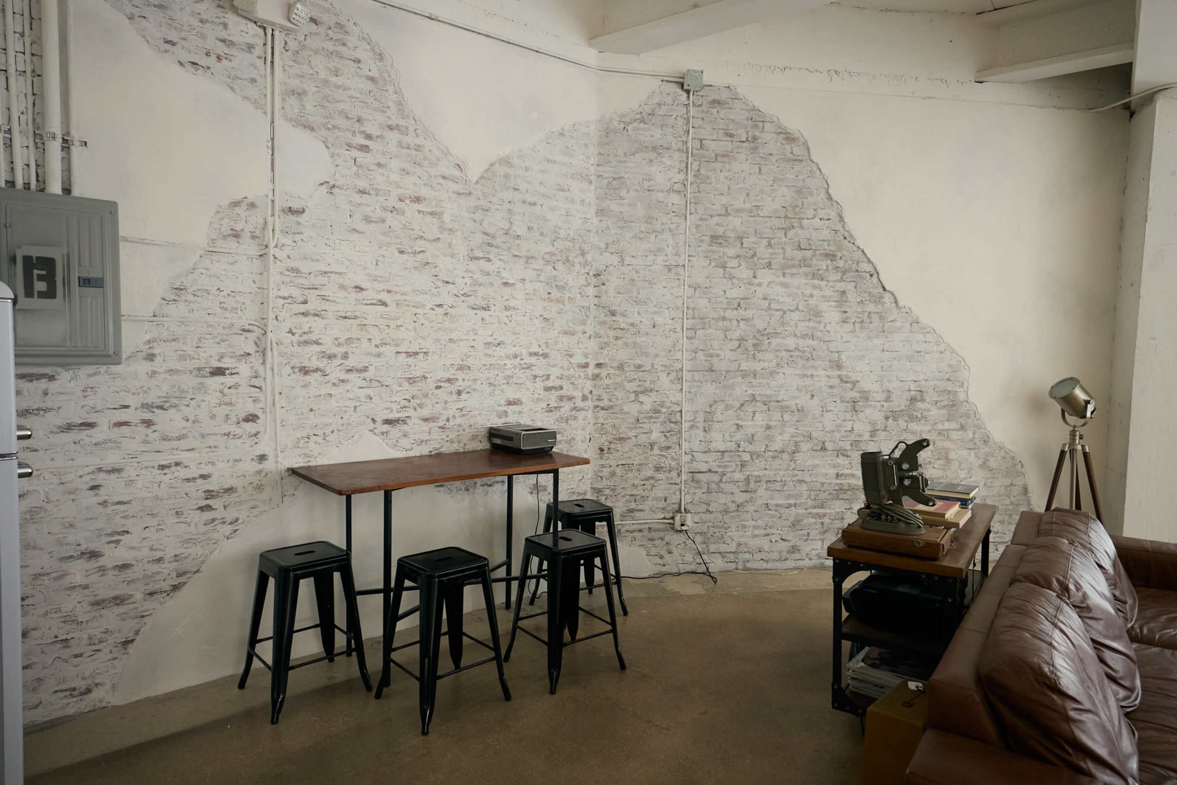 The image shows a room with an exposed brick wall, a wooden table, several stools, a vintage typewriter, and a bronze lamp, set in a contemporary interior.