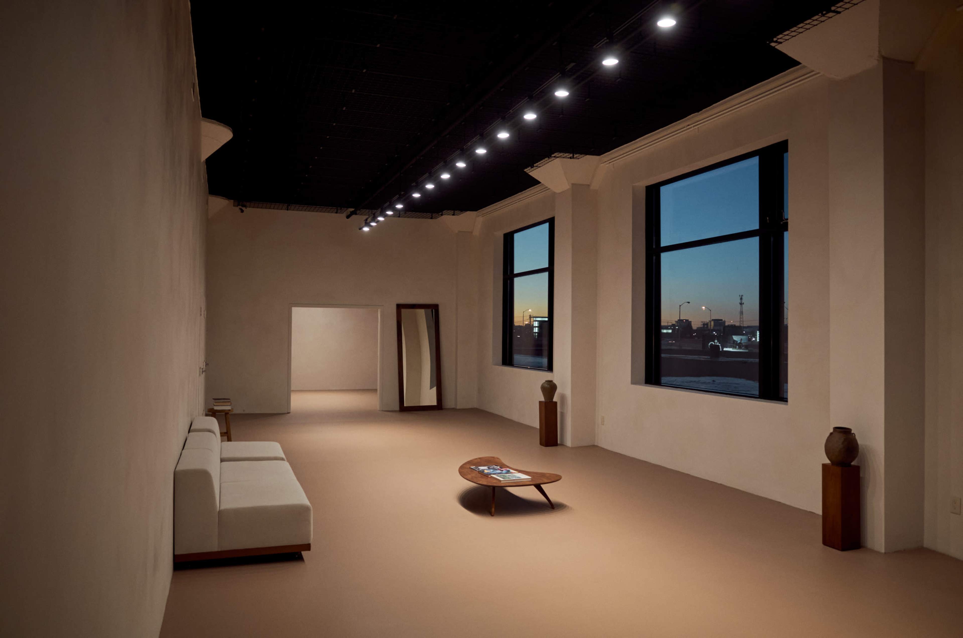 The image shows a minimalist interior space with a light-colored floor, two large windows, a white sofa, a wooden coffee table, and a mirror on the wall.