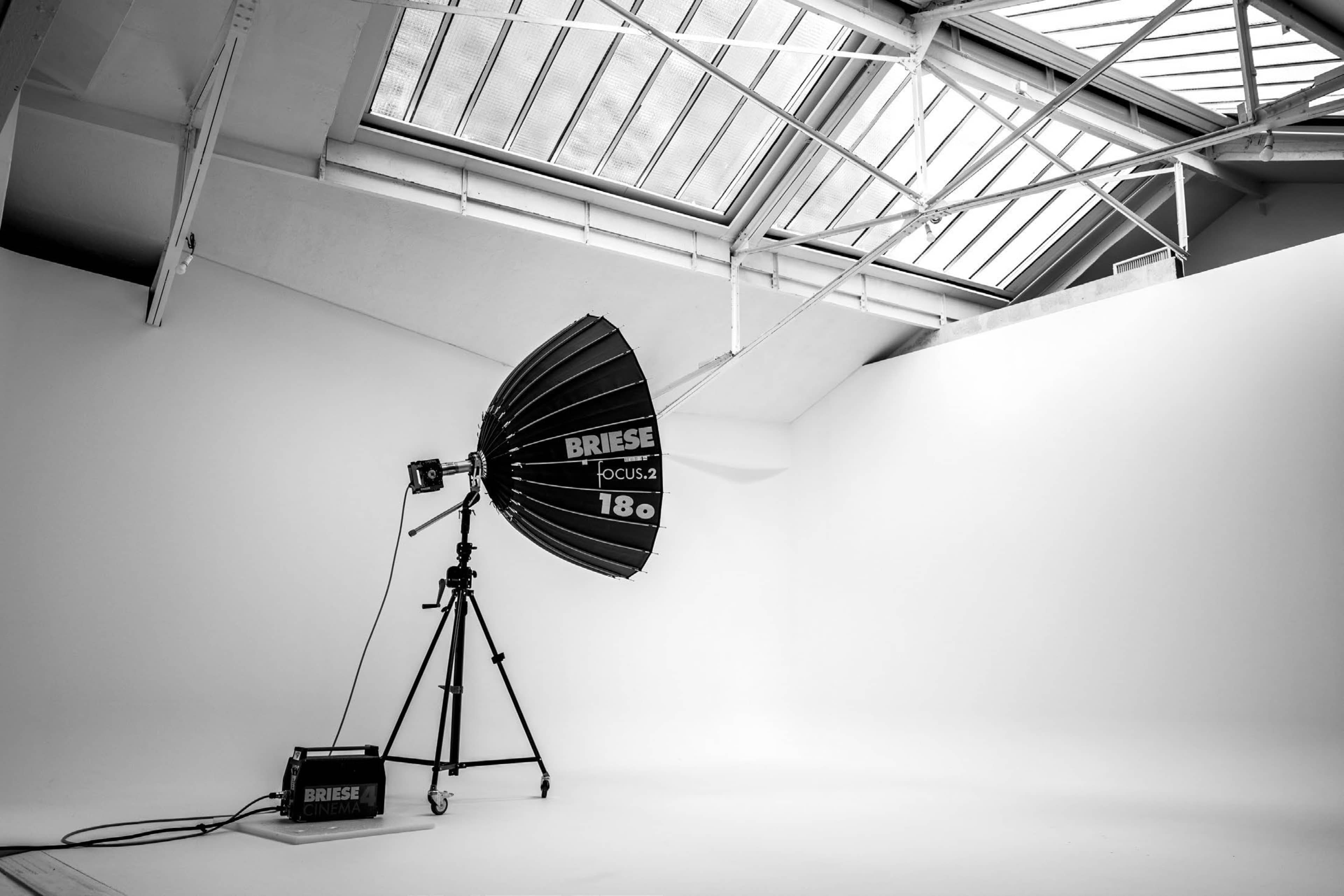 A photography studio features a large softbox light mounted on a tripod, with a backdrop of white walls and a skylight above.